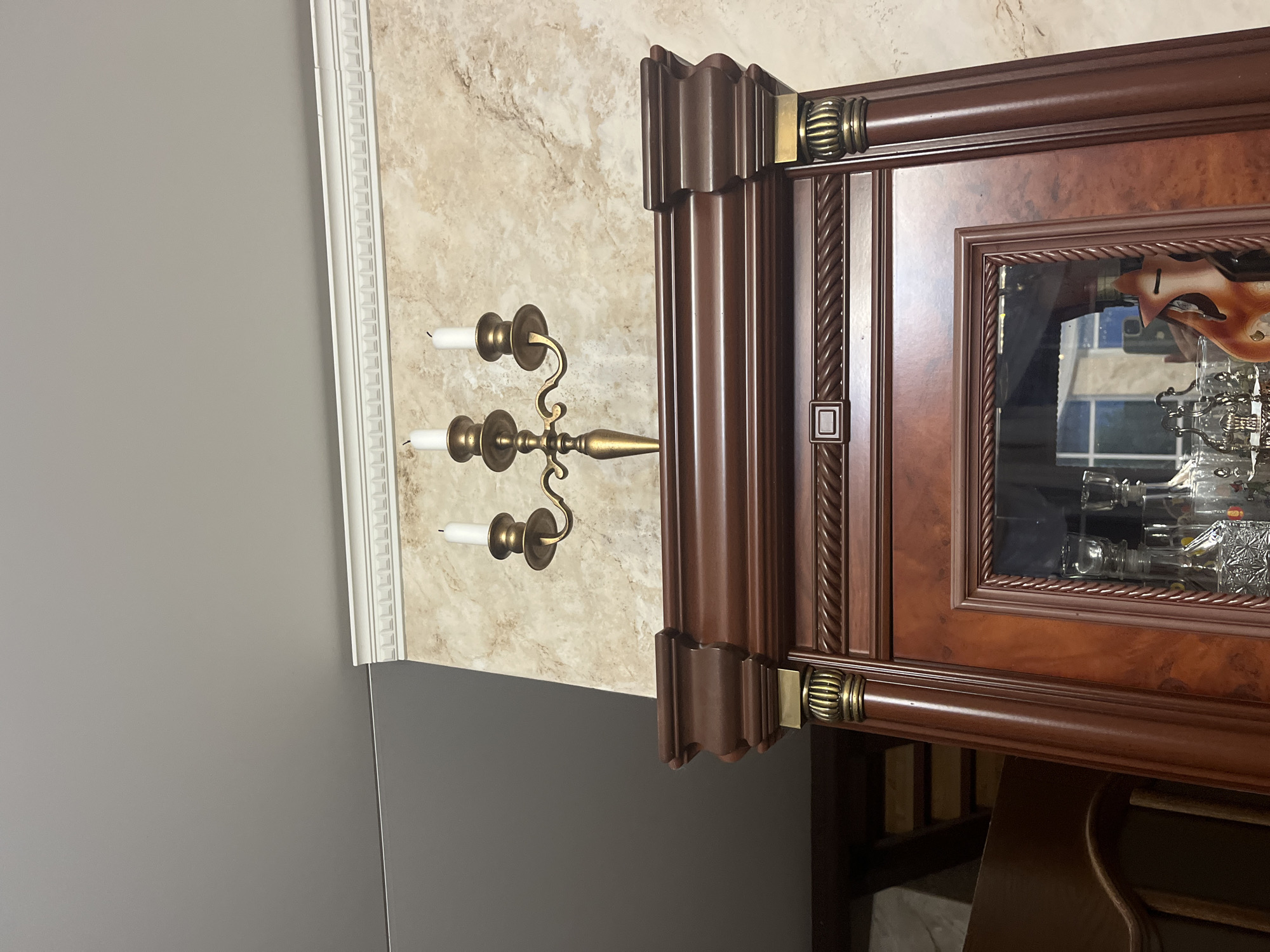 The image shows a close-up of the top of a wooden cabinet, with a candelabra holding three unlit white candles positioned on top. The cabinet has ornate detailing, including a rope-like trim and what appear to be decorative finials with gold accents. A mirrored section is visible at the front of the cabinet, reflecting some bottles and a stylized wooden fox decoration. The background features a textured, marble-like wallpaper. To the left of the cabinet, a portion of a gray ceiling is visible, with a white decorative molding strip running along the top edge of the wall. The image is taken indoors, and there are no people or animals present. The specific location within Vladimirovka, Russia, cannot be determined from the visual information provided.