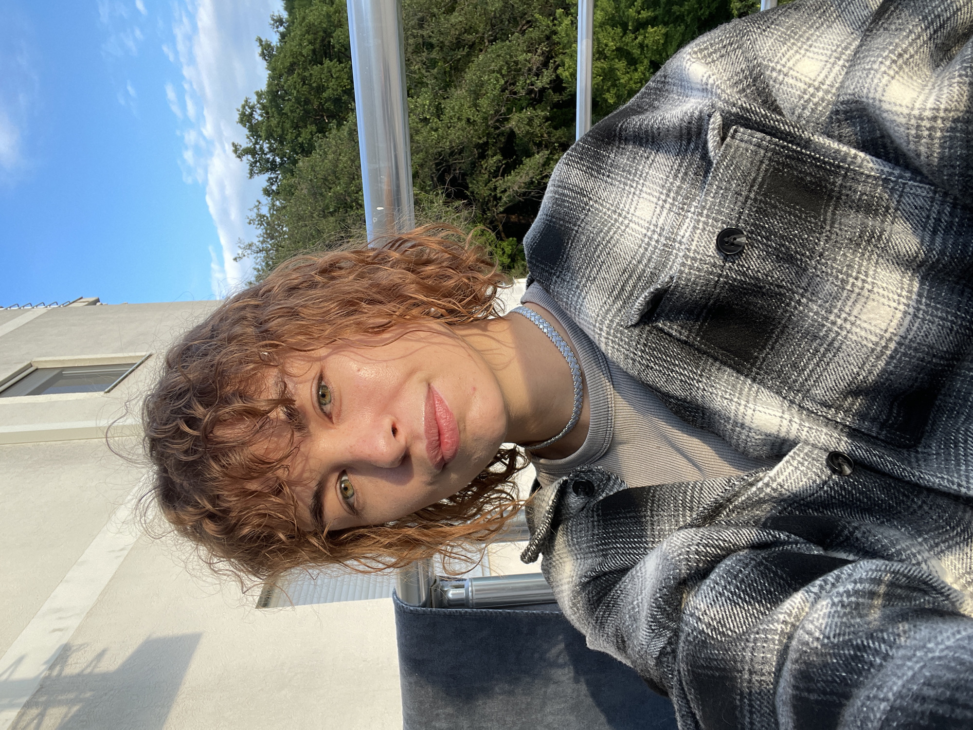A selfie of a young woman with reddish-brown curly hair, wearing a grey-and-black plaid shirt over a grey top and a silver chain necklace. She has green eyes and is looking at the camera. The background shows a bright sky with clouds, green trees, and a building. The location appears to be a balcony or terrace with a metal railing. Based on the background elements, the location is likely Rafailovići, Montenegro.