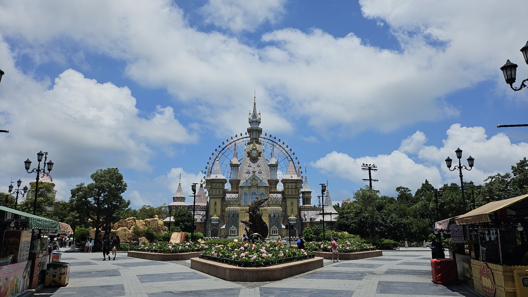 The image depicts a scene at Sun World Hon Thom Nature Park in Phú Quốc, Vietnam. In the center, a large, ornate castle-like structure with multiple spires and a Ferris wheel integrated into its design dominates the background. In front of the castle, there is a sculpted figure, possibly a dragon, surrounded by flower beds filled with pink and white blossoms. The foreground shows a paved walkway with people strolling and a few food stalls or kiosks on either side. Lampposts with multiple globes are visible throughout the scene, and lush green trees frame the background. The sky is bright blue with scattered white clouds, suggesting it is daytime and fair weather.