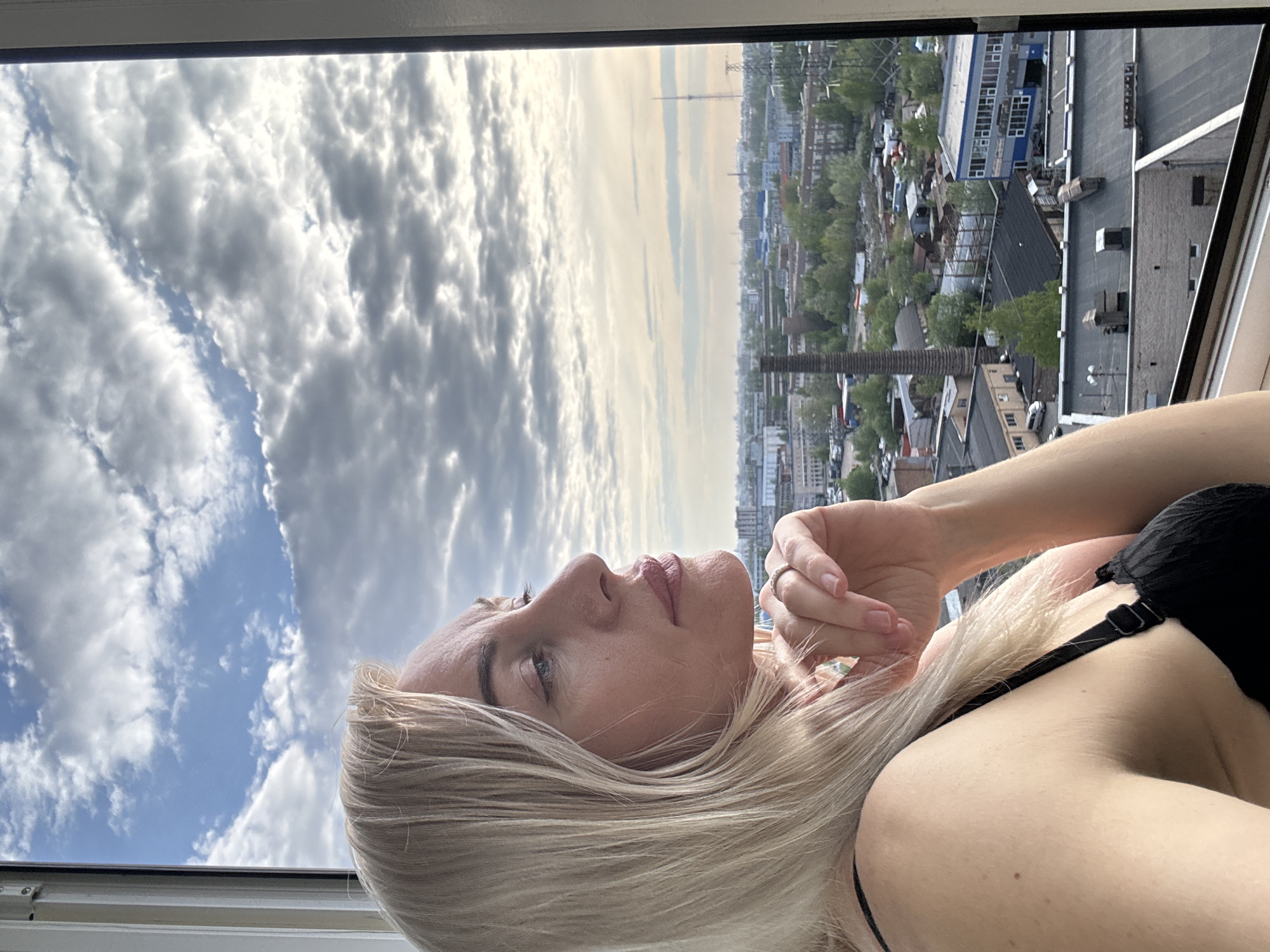 A photograph shows a fair-skinned woman with blonde hair lying horizontally near a window. She wears a black bra. Her head is tilted upward, and she gazes toward the sky, her hand near her face. A cityscape stretches in the background, featuring an array of buildings, some with visible rooftops and varying heights, interspersed with greenery. The sky is filled with patchy clouds. In the distance, a tall, thin structure rises above the buildings. The setting appears to be a window overlooking an urban area within the city of Unknown, Unknown.