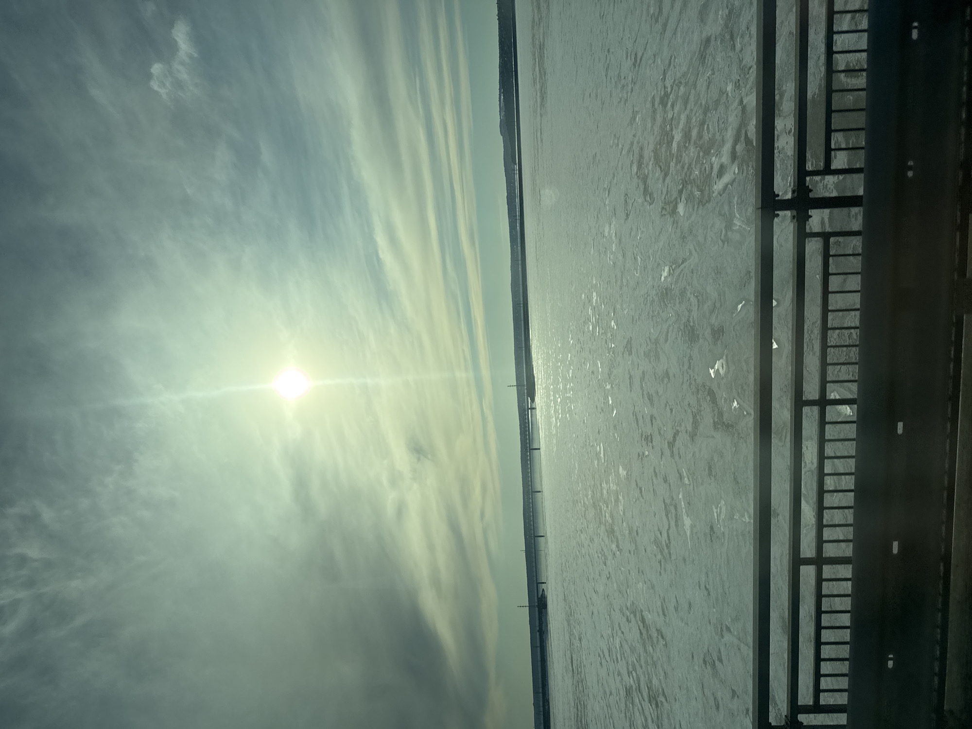 The image shows a frozen body of water next to a metal structure resembling a bridge or platform railing. The sky is bright with the sun shining through clouds, which are streaked in a linear fashion. In the distance, there is a darker band that could be the shoreline or a distant treeline. Given the location of Пальники, Russia, it is likely a frozen river or lake, observed from a bridge or an elevated structure near the water.