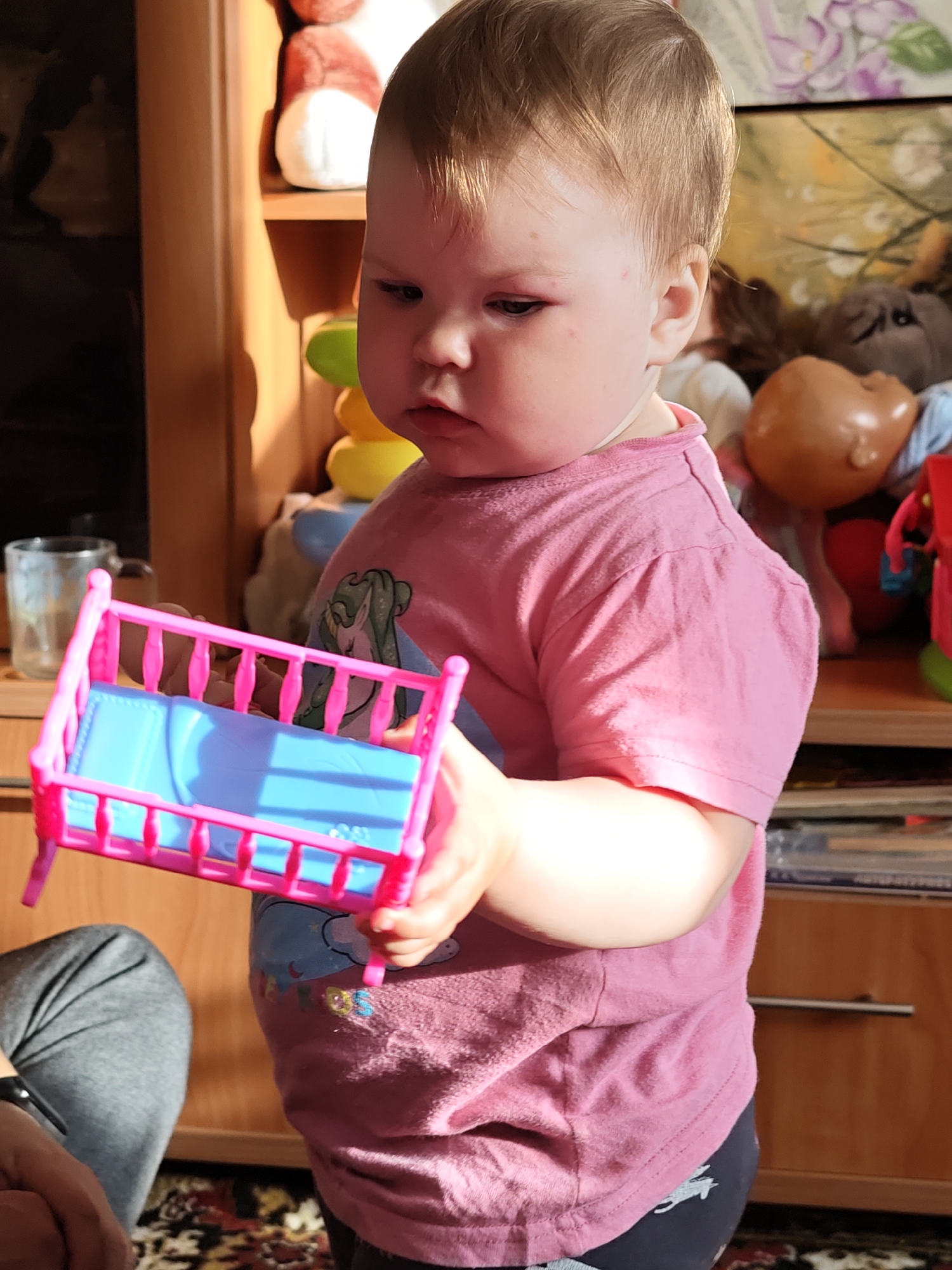 A toddler in Уральское, Russia, wearing a pink shirt and dark bottoms, holds a small pink and blue toy bed. The background includes a wooden cabinet with toys and other items on the shelves. The scene is illuminated by natural sunlight.