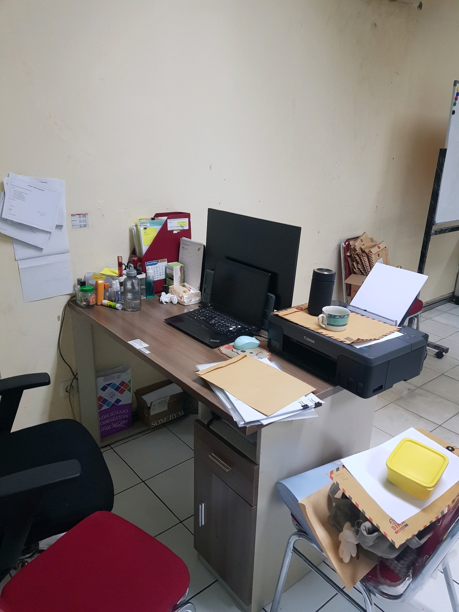 The image depicts a cluttered office desk area. On the desk, a black computer monitor is positioned behind a laptop. To the right of the laptop, a black Canon printer is present, topped with a coffee mug and some envelopes. A dark cylindrical object, possibly a thermos, stands behind the mug. To the left of the monitor, a red folder organizer holds several documents. Various office supplies, including pens, bottles, and a small jar, are scattered across the desk. A roll of paper, labeled "BRIGHT WHITE OFFICE PAPER," is visible under the desk on the left. Two office chairs are present: one black chair to the left and a red chair partially visible in the lower right. The walls appear to be painted a light color. In the background, a whiteboard stands on a mobile frame. The overall impression is of a functional but somewhat disorganized workspace. The location is described as Central Jakarta, Indonesia, suggesting an urban office environment.