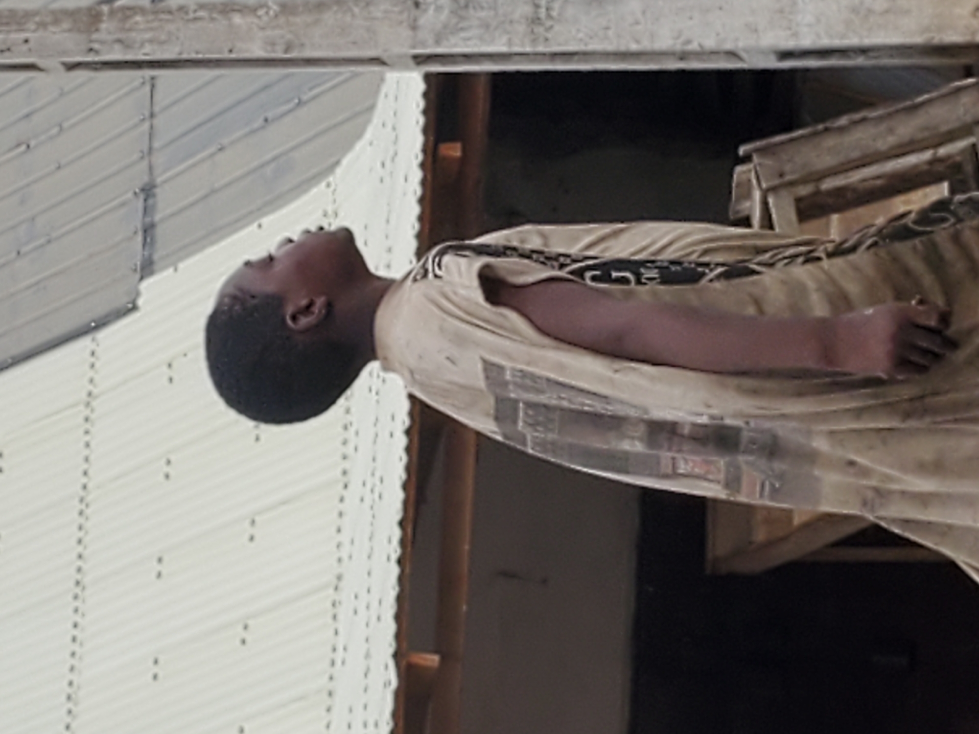 An African boy lies on a wooden table inside a corrugated iron building in Mararaba, Nigeria. He is wearing a beige t-shirt and pants. He is lying on his back.