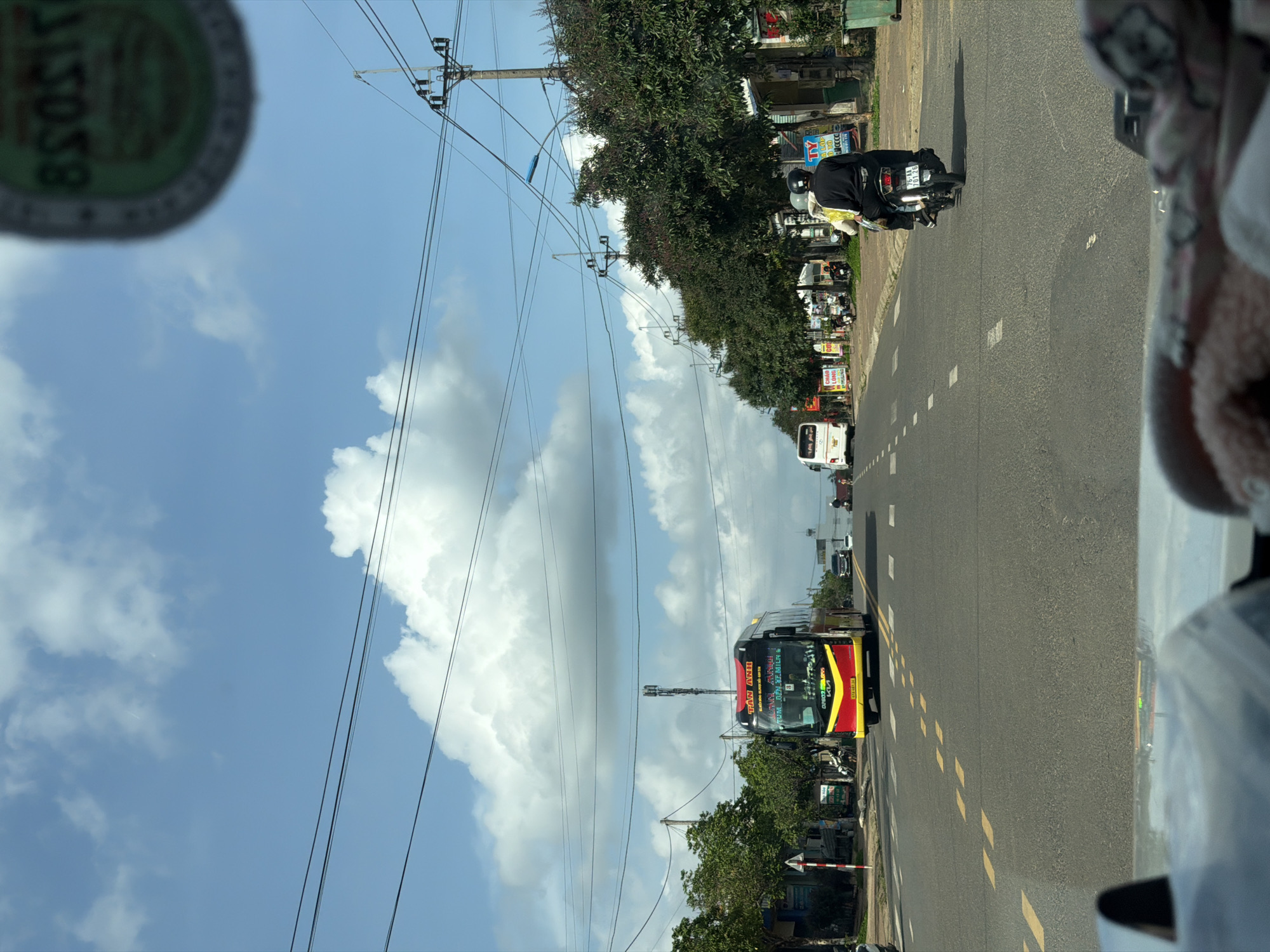 The image depicts a view from inside a vehicle, possibly a car, traveling on a road in Măng Đen, Vietnam. A red and yellow bus with the words "TAN ANH" written on the front is visible on the road, heading in the same direction as the vehicle. A motorbike with two people on it is seen further up the road. Utility poles and wires line the side of the road. The sky is blue with white clouds. Trees and buildings are visible on the side of the road.