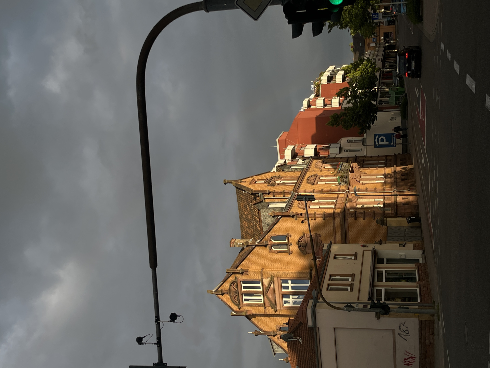 The image captures a street scene in Landau in der Pfalz, Germany, under a dark, overcast sky. A street lamp dominates the foreground on the left side. Traffic lights show a green signal. A street stretches into the distance on the right. A black car is traveling along the road. The buildings are a mix of red and yellow brick structures. The building closest to the viewer features windows and roof details. Graffiti is visible on the building in the bottom right corner. The image suggests a late evening or early morning setting.