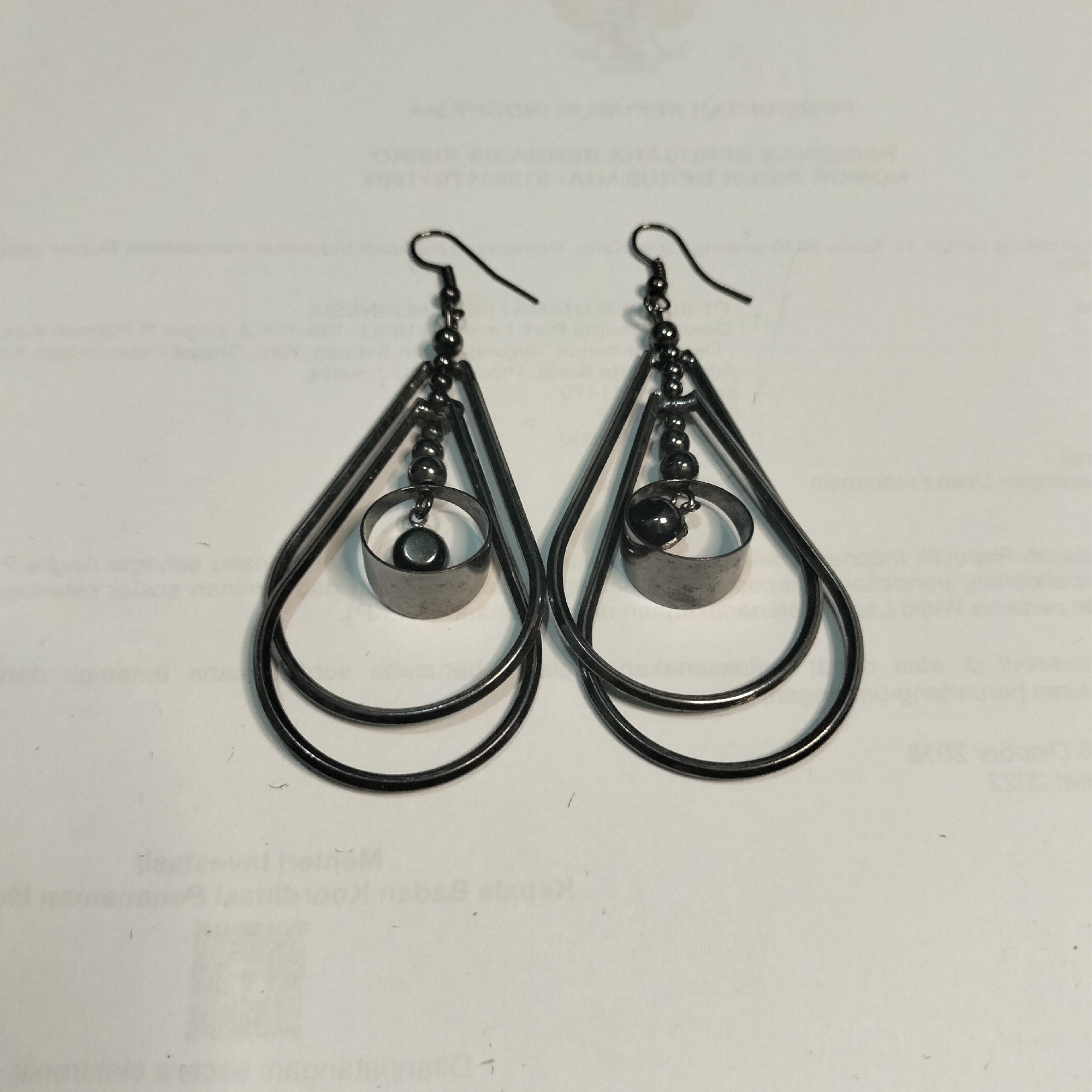 A pair of dark grey teardrop-shaped earrings are displayed against a white background with visible text printed on it. The earrings are made of thin metal wire, forming two concentric teardrop shapes. Each earring has a hook for attachment at the top, followed by a string of small, dark beads. Below the beads, a cylindrical charm hangs, with a small, dark sphere attached to its center. The background appears to be a document, possibly with Indonesian text, suggesting the location in Tangerang, Indonesia. There are no people or actions depicted in the image, only the stationary earrings.