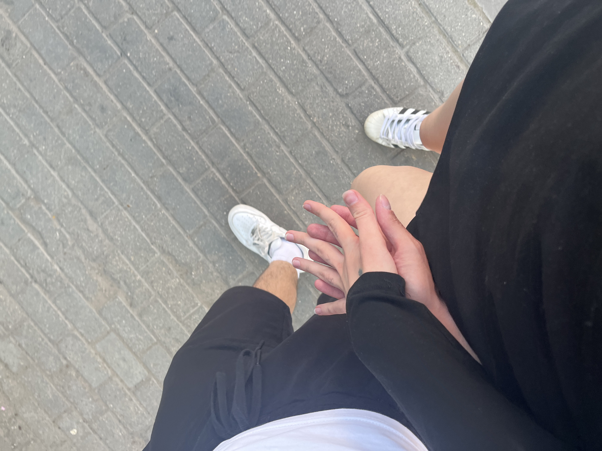 An overhead view captures two individuals holding hands while walking on a grey paved surface. The ground is composed of rectangular pavers laid in a diagonal pattern. The person on the left wears a white short-sleeved top, black shorts, white athletic socks, and a white sneaker, with a bare leg visible. The person on the right wears a black long-sleeved top, a black skirt, and a white Adidas sneaker with black stripes, with a bare leg visible. Their hands are intertwined, palms facing each other, with a small tattoo visible on the right person's wrist area. The scene is outdoors in the city of Unknown, Unknown.