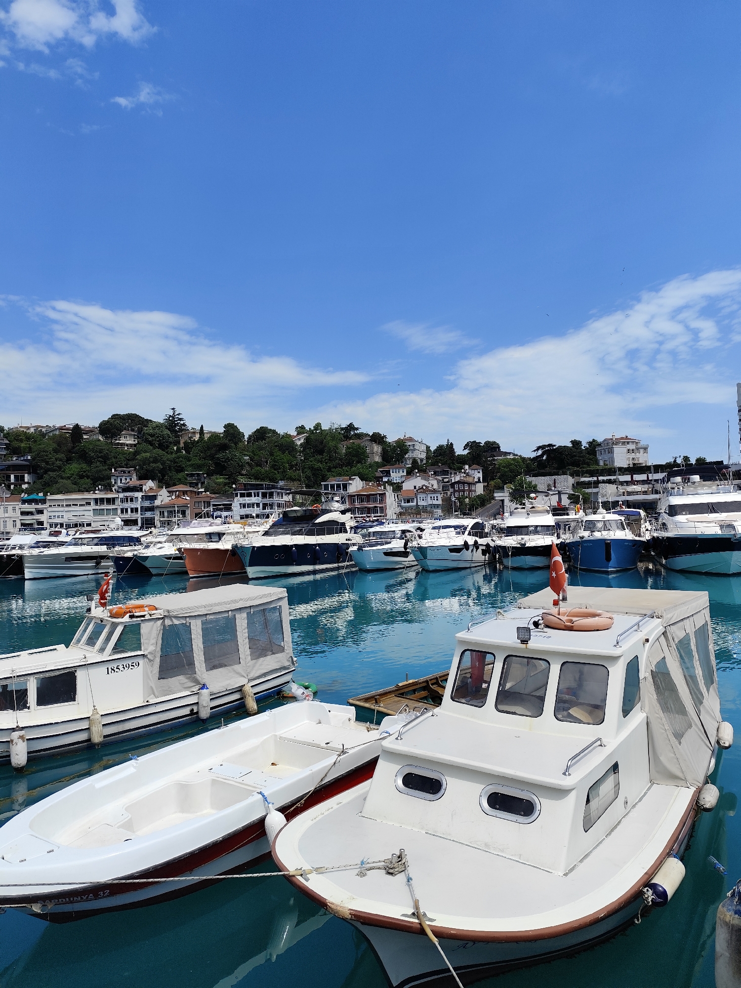 This image captures a serene marina scene in Sarıyer, Turkey, on a clear, sunny day. The foreground features several boats, with the background showcasing a bustling harbor flanked by a lush green hillside dotted with residential buildings.
In the immediate foreground, to the left, is a white boat with a grey canvas canopy, bearing the identification number "1853959" on its side. Adjacent to it, a smaller white dinghy or rowboat is visible, with "SARDUNYA 32" faintly inscribed on its bow. To the right, another white boat with a small cabin, porthole windows, and a grey canvas canopy is prominent, featuring an orange life buoy on its roof and a small Turkish flag proudly displayed at its stern.
Behind these foreground vessels, numerous other boats of varying sizes and colors—including white, blue, and orange yachts—are neatly docked in the marina. The water is a striking turquoise-blue, reflecting the bright sky and the boats.
The backdrop consists of a verdant hillside densely covered with trees and a mix of white and earth-toned residential buildings, some appearing quite large. The sky above is a vibrant blue, sparsely dotted with fluffy white clouds, indicating pleasant weather. No people are directly visible, contributing to the tranquil atmosphere of the scene. The overall impression is one of a peaceful, picturesque coastal town under a bright sky.