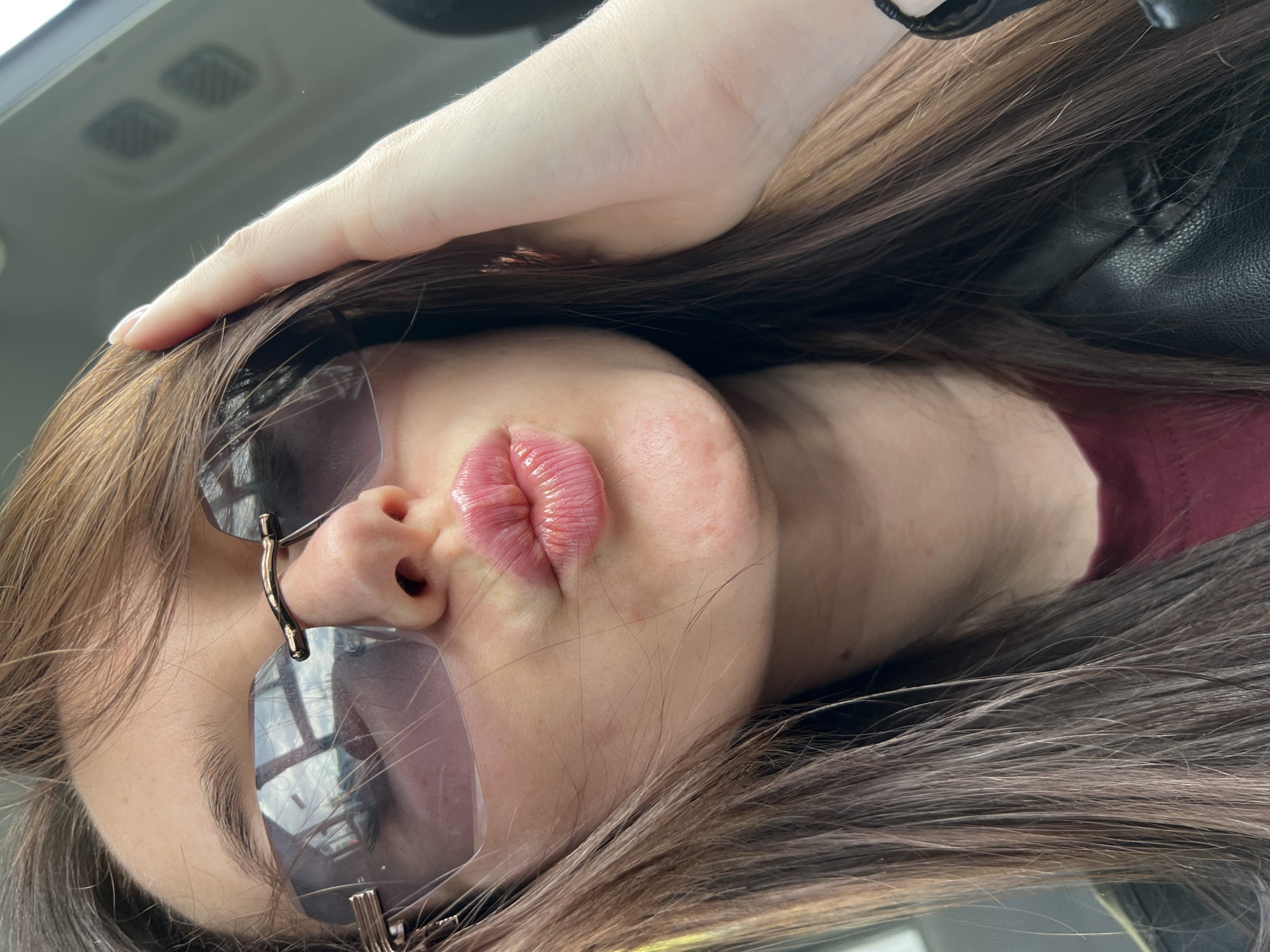 A close-up, slightly angled shot depicts a woman with long, brown hair wearing sunglasses and a black leather jacket. Her hand rests on her forehead, partially obscuring her hair. Her lips are pursed. The reflection in her sunglasses appears to show an interior setting, possibly a vehicle. The photo was taken in the city of Tyumen, Russia.
