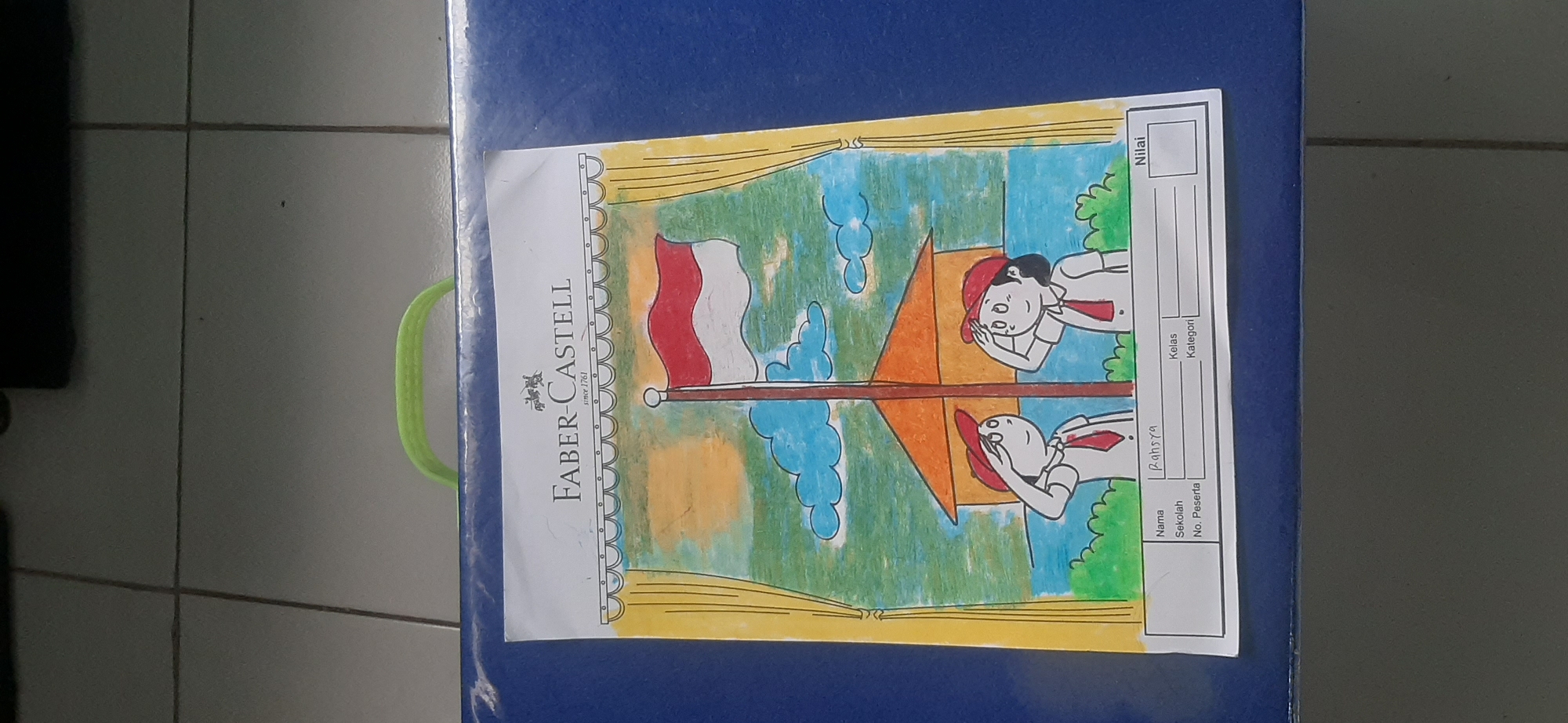 The image shows a close-up of a drawing on a piece of paper, which is placed on a blue surface. The drawing depicts two children in school uniforms saluting a flag. The flag appears to be the Indonesian flag, with red and white colors. Behind the children is a building with an orange roof, and the sky is depicted with clouds and an orange sun. The drawing is framed by yellow curtains on either side. At the bottom of the drawing, there is a section with lines and boxes for information such as "Nama" (Name), "Sekolah" (School), "No. Peserta" (Participant Number), "Kelas" (Class), "Kategori" (Category), and "Nilai" (Score). The name "Rahsya" is written in the "Nama" field. The text "Faber-Castell since 1761" is visible at the top of the paper, indicating the brand of art supplies used. The blue surface appears to be part of a case or bag. The background shows a tiled floor. The image does not contain any specific elements that would allow for the identification of the city of Jambi, Indonesia.