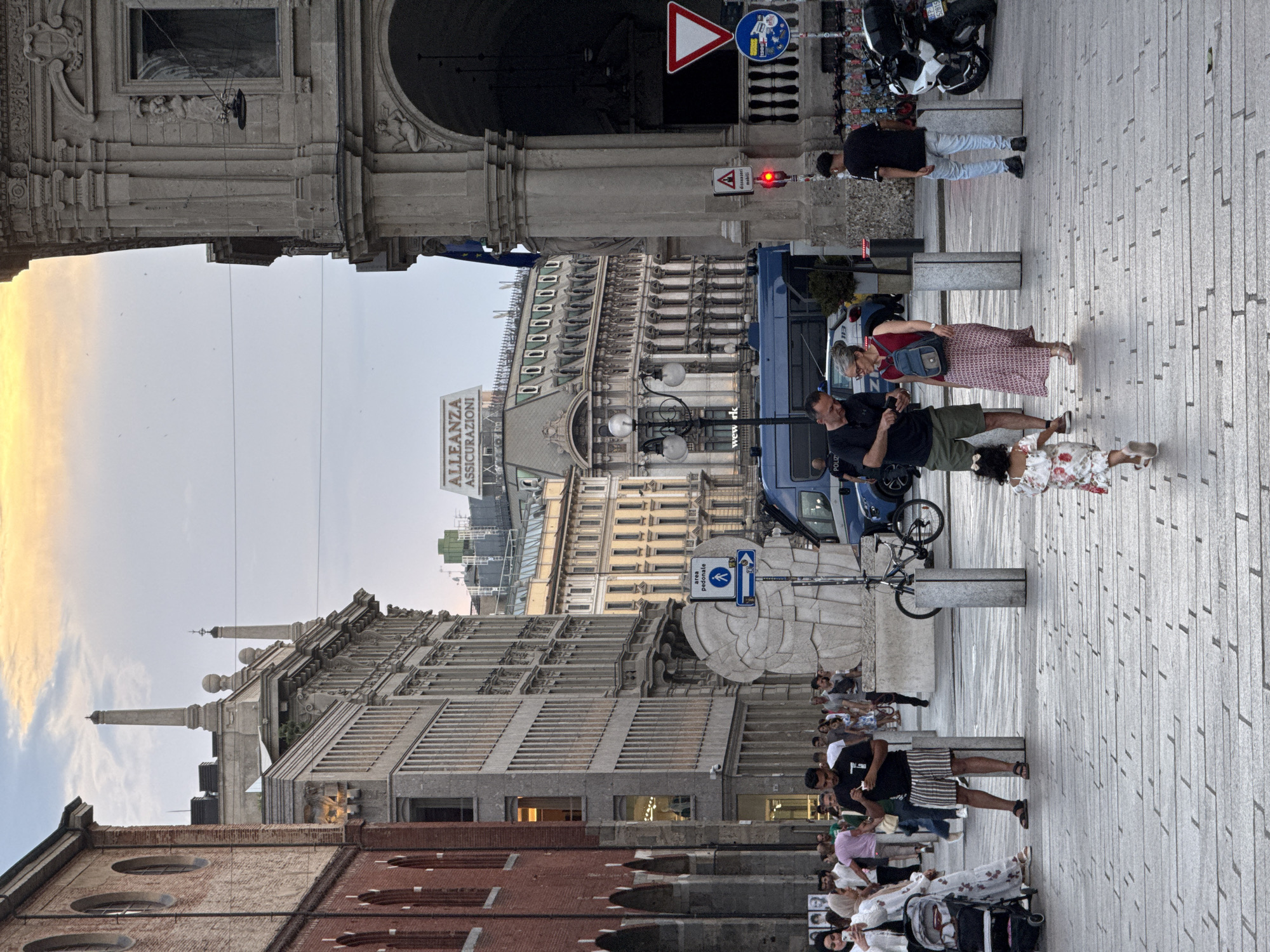 The image captures a street scene in Milan, Italy, likely in Piazza del Duomo or a nearby square, given the architectural style. The time of day appears to be late afternoon or early evening, with warm sunlight illuminating the sky and casting long shadows.
In the foreground, a cobbled pedestrian street is visible, with several people walking. A young girl in a floral dress is running towards the right of the frame. To her left, a couple is standing and interacting with a smartphone. The man, wearing a black t-shirt and striped shorts, is holding the phone, while the woman, in a red top and patterned skirt, has a backpack. Further left, a woman with a stroller is seen amidst a group of pedestrians.
In the mid-ground, a distinctive large sculpture, appearing to be made of stone or concrete and resembling a stylized head or shell, is prominent. A blue police van is parked behind it, and a bicycle is leaning against a bollard. Traffic signs, including a pedestrian crossing sign, are visible.
The background features grand, historic buildings with ornate architectural details, characteristic of Milan's urban landscape. An "ALLEANZA ASSICURAZIONI" sign is visible on a building in the distance, suggesting a commercial area. The sky is partly cloudy with hints of sunset colors. On the far right, motorcycles are parked, indicating a public transportation or parking area. The overall atmosphere is that of a busy urban public space.