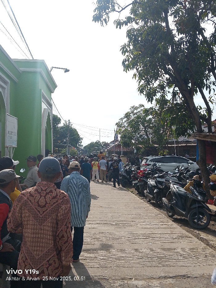 A daytime scene shows a crowded street lined with parked motorbikes and trees. People are walking in the street, some wearing casual clothes, others in shirts and trousers. A green building is on the left side. The street is made of concrete and has visible lines across it. The sky is bright and clear. The photo was taken with a Vivo Y19s, as indicated in the bottom left corner.