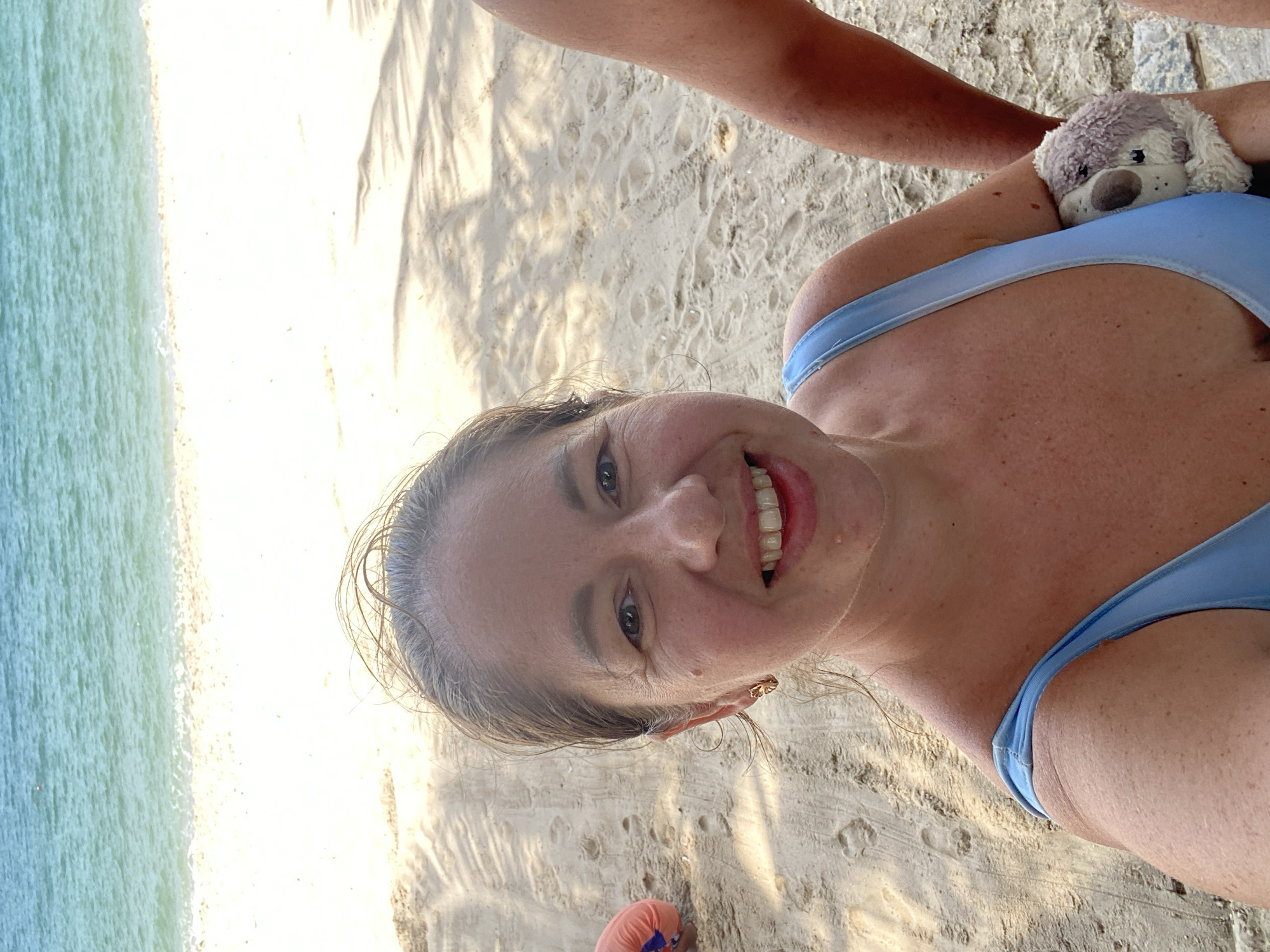 The image shows a woman in a light blue swimsuit on a beach. She is smiling and looking at the camera. She is holding a small stuffed animal in her arms. In the background, there is the ocean and a sandy beach with palm tree shadows. To the left of the woman, a person wearing an orange garment is partially visible. To the right of the woman, another person's arm is visible. The location is identified as Phu Thuy Ward, Vietnam.