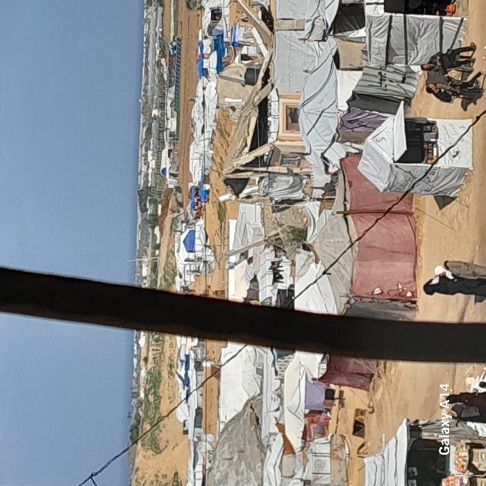 The image presents an expansive, elevated view of a densely populated makeshift encampment situated in Khan Yunis, Palestinian Territory. The terrain is largely sandy and uneven, transitioning to low hills with sparse vegetation in the background, beneath a clear, light blue sky that meets a hazy horizon, possibly indicating a distant body of water.
The encampment consists of countless temporary shelters, predominantly constructed from white, grey, blue, and maroon tarpaulins draped over frames or existing damaged structures. Several shelters show signs of wear, including collapsed sections and exposed building materials, while others appear to be simple tent arrangements. Laundry hangs on lines outside some of the structures.
In the foreground, a dark, diagonal beam obstructs part of the view, with wires visible crisscrossing the frame. A small, white, tent-like kiosk with an open counter displaying goods stands on the dirt ground. Two male figures are seated on chairs to the right of this kiosk. Further into the mid-ground, two figures, one in dark attire including an abaya, stand near maroon-colored tents. Numerous other indistinct figures are visible throughout the sprawling camp, engaged in various activities. The bottom-left corner of the image contains a "Galaxy A14" watermark.