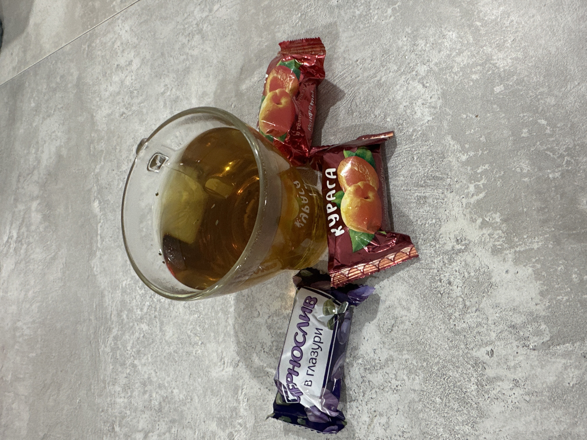 In an indoor setting, likely a kitchen or living room, on a textured grey surface resembling a countertop or table, sits a clear glass mug filled with amber-colored liquid, presumably tea. The mug has a handle on the right side. Adjacent to the mug are three individually wrapped candies.
On the left, a candy wrapper is predominantly purple and white, adorned with images of blueberries or plums. The text on this wrapper reads "ЧЕРНОСЛИВ В ГЛАЗУРИ" which translates to "Prunes in glaze".
In the center and slightly forward, another candy wrapper, primarily maroon and gold, features an image of apricots. The text on this wrapper says "КУРАГА" meaning "Dried apricots".
To the right of the mug and slightly behind the apricot candy, a third candy wrapper, also maroon and gold, displays a similar image of apricots and the text "В тёмной глазури Конфеты" which translates to "In dark glaze Candies".
The overall scene is a still life composition of a beverage and sweets. The specific location of Chaikovsky, Russia, is not visually determinable from the provided image itself.
