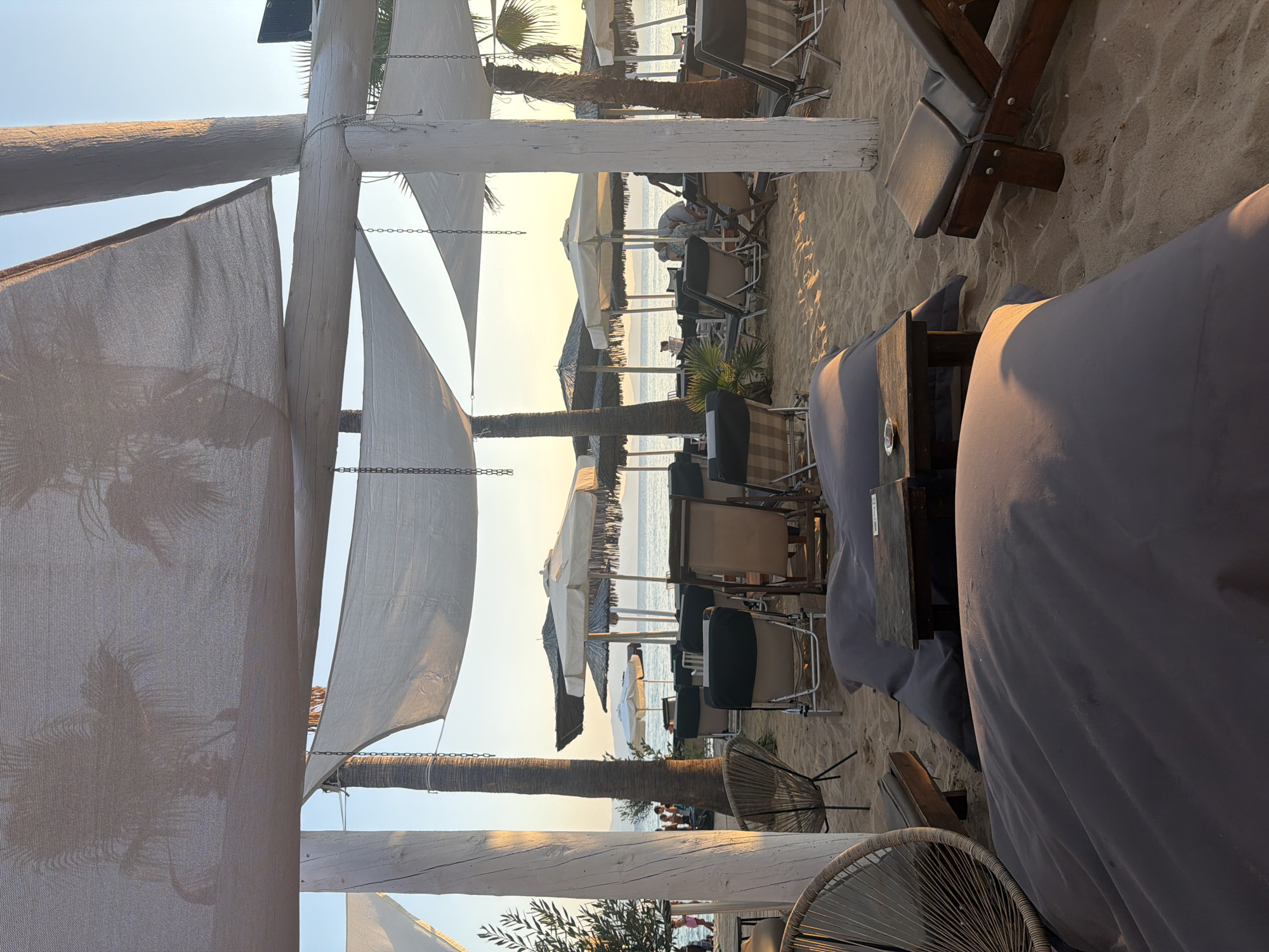 The image depicts a beachfront seating area at a cafe or restaurant in Ofrynio Beach, Greece. Large white fabric canopies, supported by white wooden posts and chains, provide shade over a sandy area furnished with lounge chairs, beanbag chairs, and low tables. Palm trees and other foliage are visible behind the seating. In the background, beach umbrellas are set up on the sand, and people can be seen walking near the water's edge. The sea is calm, and the sky is a clear, pale blue, suggesting either early morning or late afternoon. The lighting indicates a warm, sunny day.