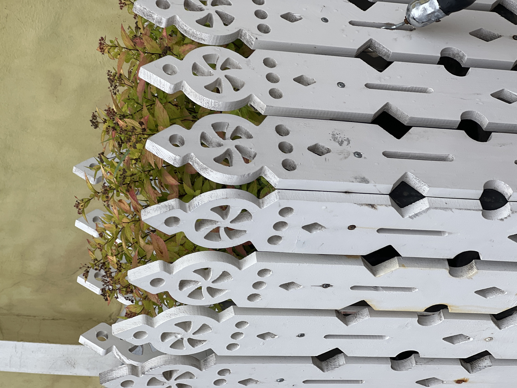 The image shows a close-up, eye-level view of a white, decorative wooden fence with a green and red-leafed plant visible behind it. The fence has intricate cut-out designs, including circular patterns at the top of each picket and diamond shapes below. The paint on the fence appears weathered, with some areas showing signs of rust and wear. The background wall is a muted yellow color. A metallic object, possibly a tool or part of a structure, is visible on the right side of the frame, partially obscuring a section of the fence. The scene is outdoors, and the lighting suggests it is daytime.