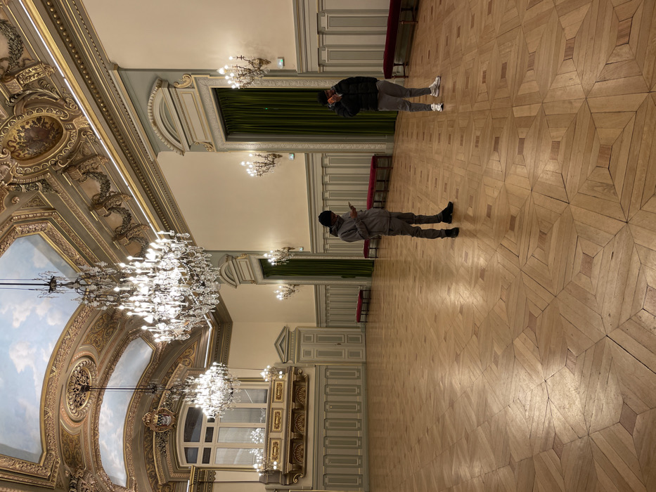 The image displays a grand, opulent hall within what appears to be a historic building, consistent with a location like Paris, France. The hall boasts high ceilings elaborately adorned with intricate decorative moldings, gilded accents, and painted panels that depict a sky with clouds. Multiple large, sparkling crystal chandeliers hang from the ceiling, illuminating the space with bright artificial light.
The walls are a light color, featuring classical-style paneling and pilasters, with dark green velvet curtains visible in recessed window or alcove areas. Along the walls, there are simple, long, dark red benches. The floor is a prominent feature, crafted from light-colored wood laid in a complex and visually striking geometric parquet pattern.
Two men are present in the spacious hall. In the foreground, slightly to the left, a man wearing a light grey hooded sweatshirt, matching trousers, and dark shoes is holding and looking at a smartphone. Further back and to the right, another man, dressed in a dark puffer jacket, light grey trousers, and white sneakers, is also holding a smartphone, seemingly observing its screen or perhaps capturing the grandeur of the hall.
The activity suggests casual observation of the impressive surroundings, combined with engagement with personal devices. The lighting, predominantly from the chandeliers, indicates an indoor setting, possibly evening or a space that relies primarily on artificial illumination. The architectural style, scale, and rich detailing point to a venue of significant historical and cultural importance. There is no visible text in the image.