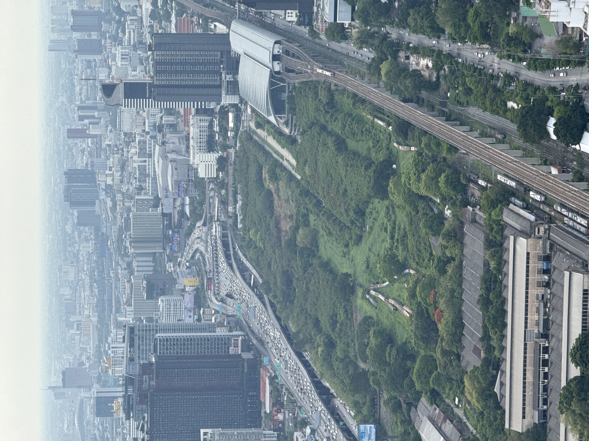 This aerial shot shows a densely populated area of Bangkok, Thailand. The image focuses on a complex infrastructure network with a mix of urban development and green spaces.
The left side shows a dense cityscape with numerous high-rise buildings, indicating a central business district or a heavily populated residential area. The buildings are varied in design and height, reflecting different architectural styles and development periods.
To the right, a large, green area forms a significant part of the landscape, suggesting a park or undeveloped land within the city limits.
Running through the image are multiple elevated highways congested with traffic, indicating heavy vehicular movement. A railway line also cuts through the scene, with a modern train station prominently visible. The station has a distinctive, curved architectural design.
The overall scene depicts a city with a blend of modernity and nature, where transportation networks are a crucial element in managing the urban sprawl.