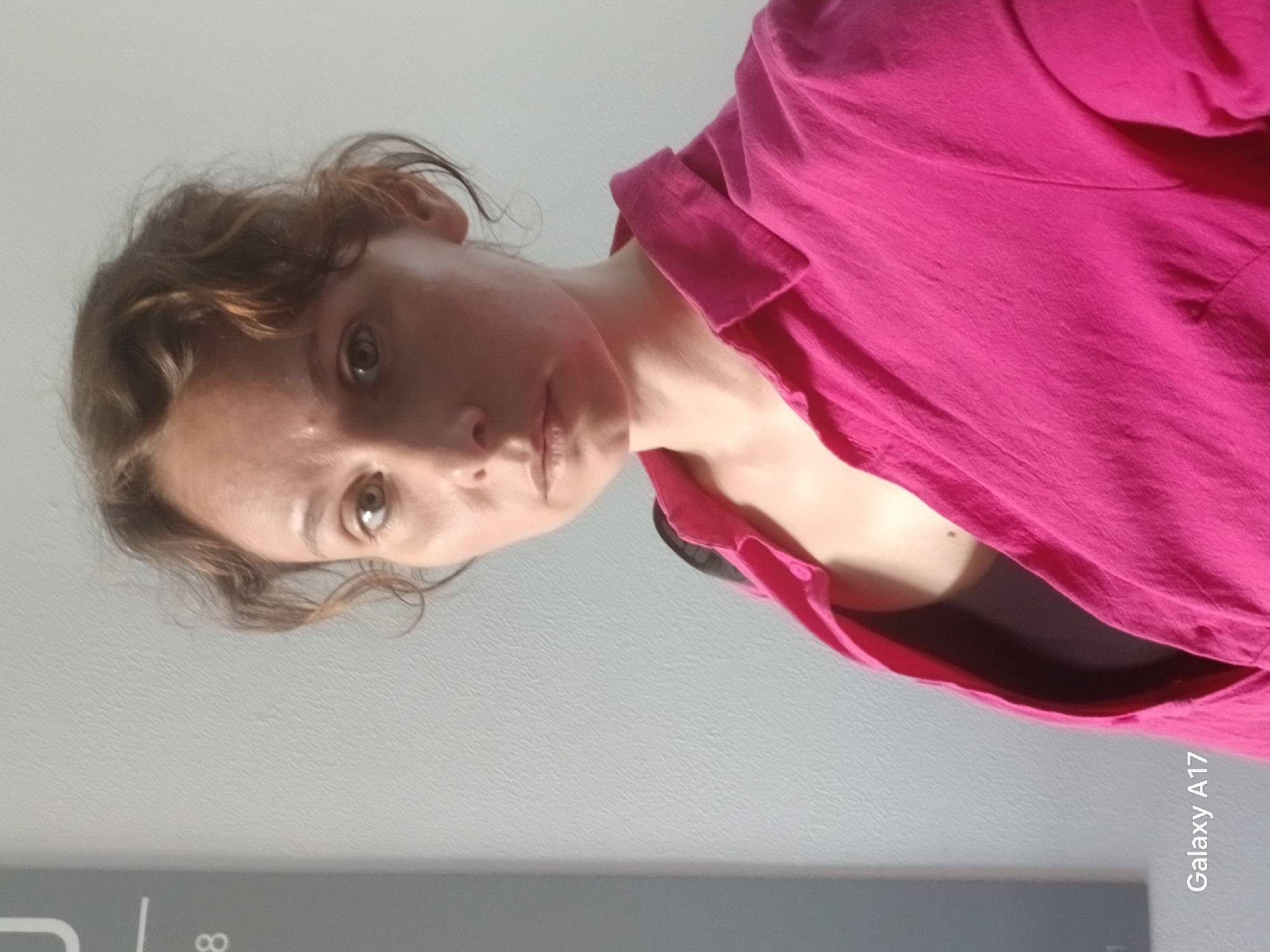 A woman with light brown hair and brown eyes is facing the camera. She is wearing a bright pink collared shirt over a dark undershirt. The background is a plain gray surface, likely a wall or floor. The phone model "Galaxy A17" is visible in the bottom right corner. There are no indicators to determine the location in Yekaterinburg, Russia.