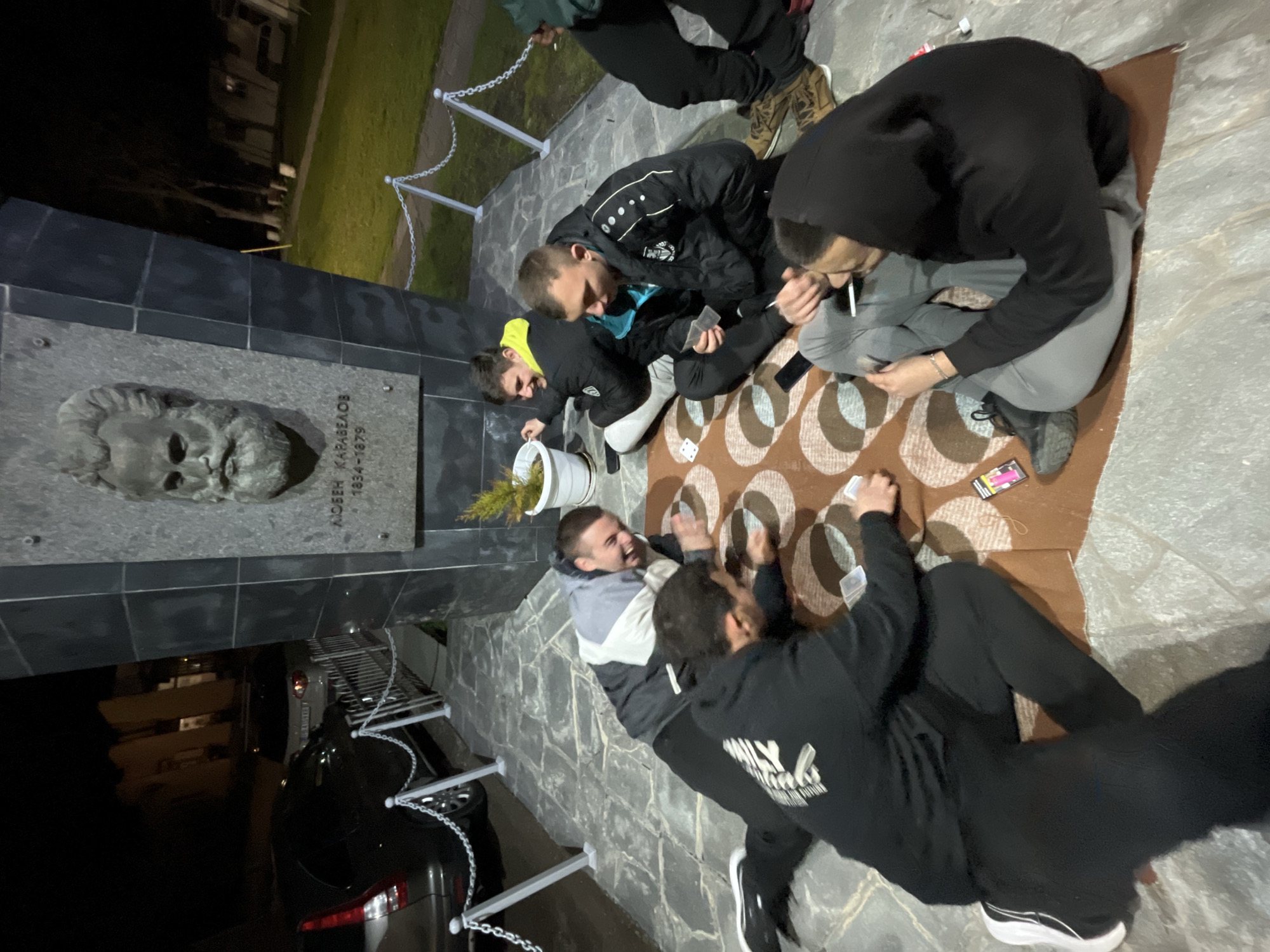 At night in Karavelovo, Bulgaria, six individuals are positioned around the monument to Lyuben Karavelov, located at an urban site, as suggested by the presence of street lighting and vehicles. One individual appears to be smoking. Some are sitting on a blanket with cards and phones near them. One individual is holding cards and yelling at another. There is a pack of cigarettes visible on the blanket.