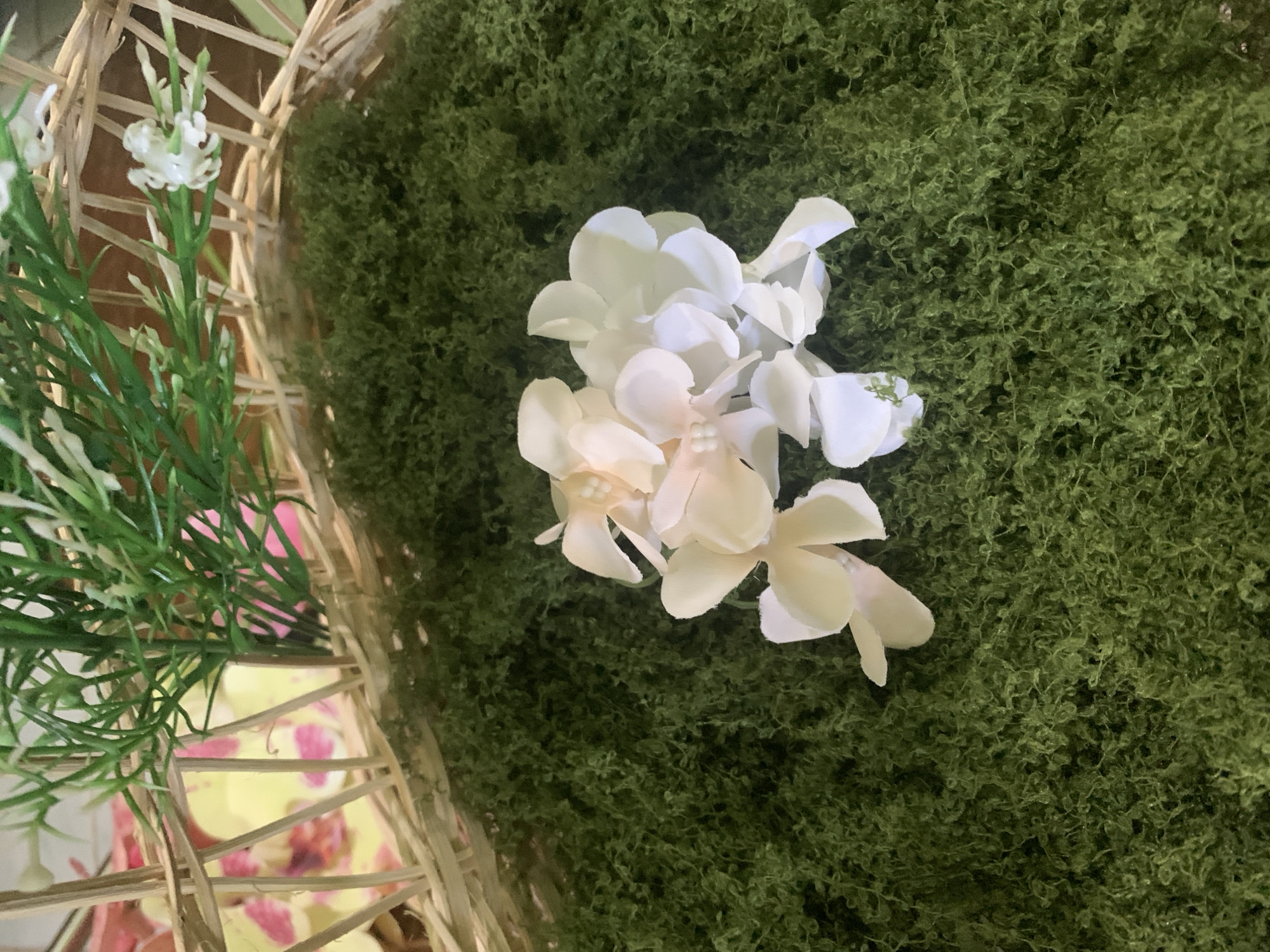 A close-up, overhead shot shows a cluster of delicate, light-colored artificial flowers resting on a bed of vibrant green moss. The flowers, some appearing off-white and others a pale peach, have multiple petals and small, bead-like centers. The moss is densely packed and has a textured, somewhat stringy appearance. Behind the moss, a portion of a woven basket is visible, with thin, light-brown reeds forming its structure. Peeking through the reeds are blurred elements of what appear to be artificial foliage, including green, grass-like stems and a few small white blossoms. In the further background, behind the basket, the indistinct shapes of pink and yellow orchid-like flowers can be seen. The scene is lit from above, casting subtle shadows. There are no people or animals present in the image, and no discernible information about a location in Kuala Pembuang, Indonesia.