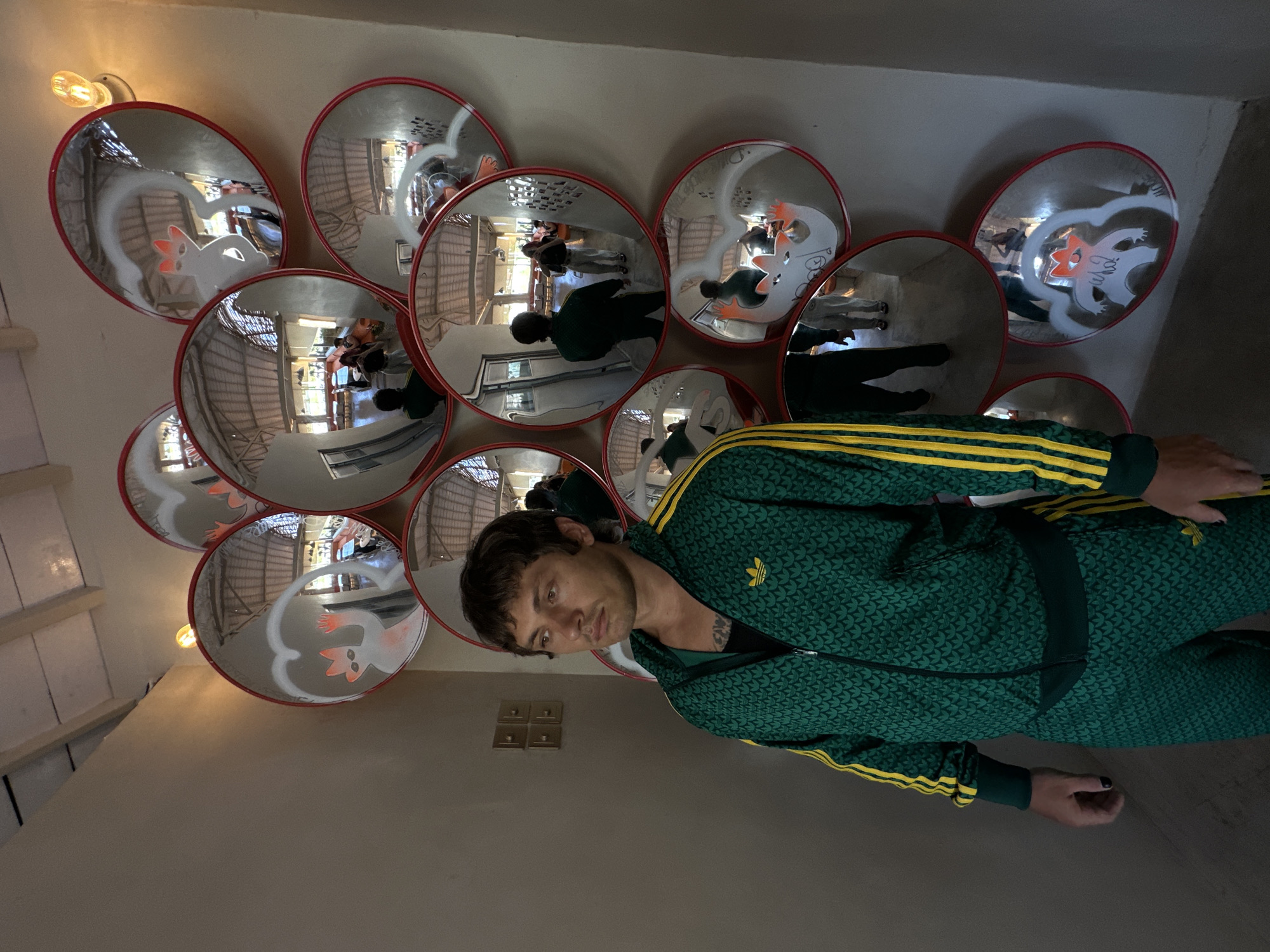 The image shows a person wearing a green Adidas tracksuit with yellow stripes, standing indoors against a beige wall decorated with a cluster of convex mirrors. The mirrors reflect a partial view of the interior, including a light source, a person, and cartoon drawings. The person has dark hair, a tattoo on their neck, and is looking at the camera. Four light switches are visible on the wall. The location is North Kuta, Indonesia.