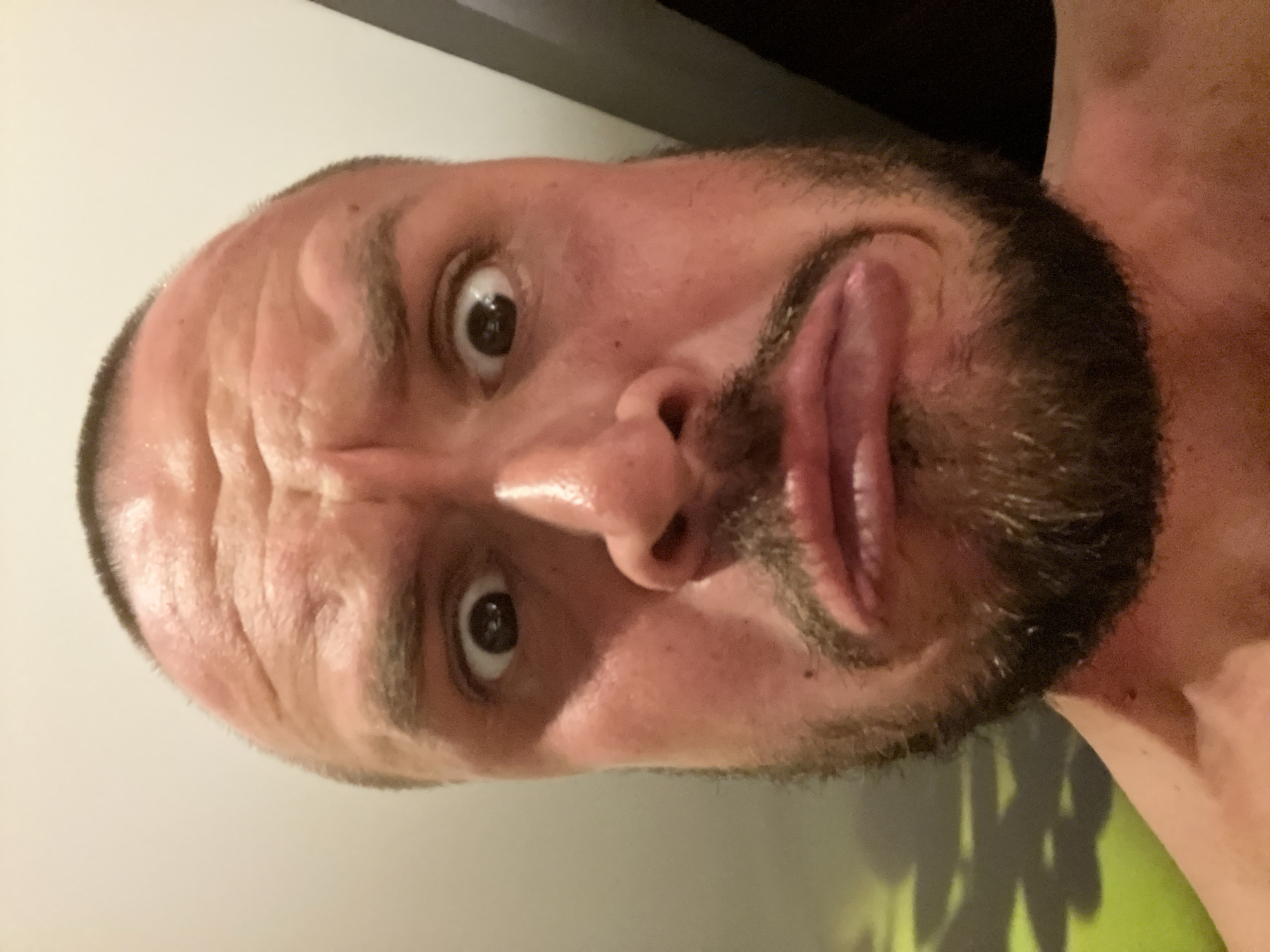 A man with a short buzz cut, brown beard, and wide, surprised eyes makes a silly face for a selfie. His eyebrows are raised and his mouth is pursed, as if blowing a raspberry. The lighting in the image is warm and intimate, casting subtle shadows on his face. In the background, a blurred green wall with a shadow pattern suggests an indoor setting. The overall impression is one of playful informality.