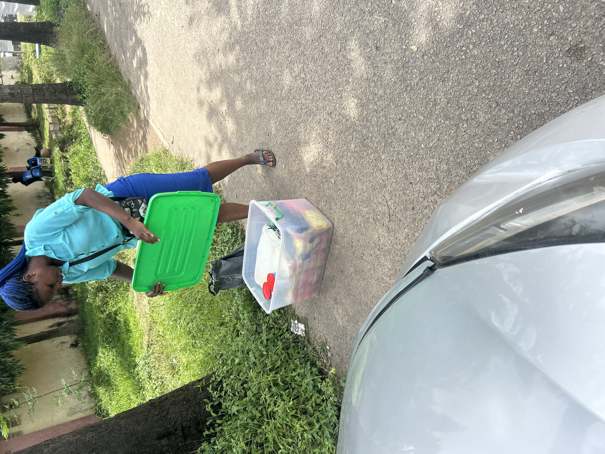 In Dutsen Alhaji, Nigeria, a woman with blue braids, wearing a turquoise blouse and blue skirt, is standing on a paved road next to a silver car. She is holding a green lid, seemingly to close a clear plastic storage bin filled with items, some of which appear to be food items in yellow and red packaging, along with white fabric. A black bag is placed on the ground near the bin. The scene is outdoors, with lush green vegetation, trees, and a building visible in the background. The lighting suggests it is daytime, with shadows cast by the trees.