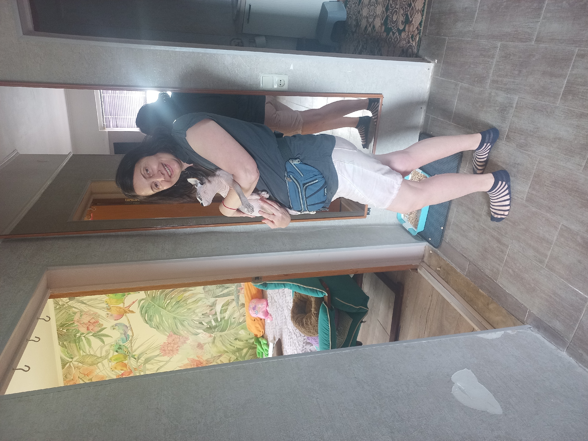 A woman holding a hairless cat stands in a doorway. The woman has long, dark hair and is wearing a dark gray shirt, white shorts, and striped socks. She has a blue fanny pack around her waist. The hairless cat is light-colored and appears to be relaxed in her arms. The doorway behind the woman leads into a room with patterned wallpaper featuring tropical plants and birds. Another doorway, visible through a mirror in the background, leads into a room with beige carpet and an area rug. The floor is tiled in a wood-grain pattern. The location is Unknown, Unknown.