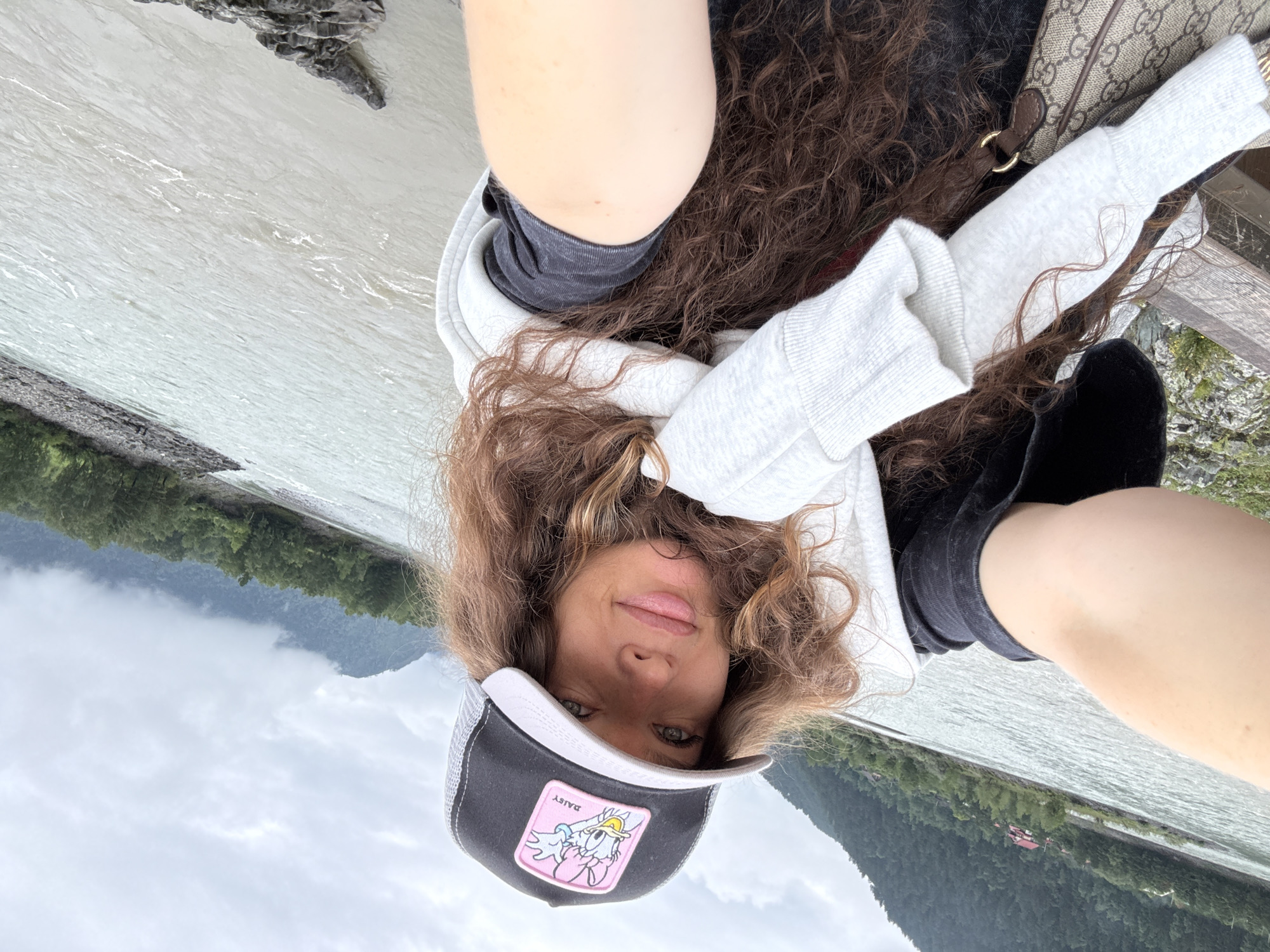 A woman is taking a selfie outdoors near a river with mountains in the background. She is wearing a black and gray trucker hat with a pink patch depicting Daisy Duck, a white sweatshirt draped around her shoulders, and dark gray t-shirt. Her long, curly brown hair is visible. She is holding her arm out to take the photo. The river is wide and appears to be flowing rapidly. The sky is overcast and the mountains are covered in green trees and some clouds. The location appears to be Elekmonar, Russia, given the presence of a river and mountainous terrain.