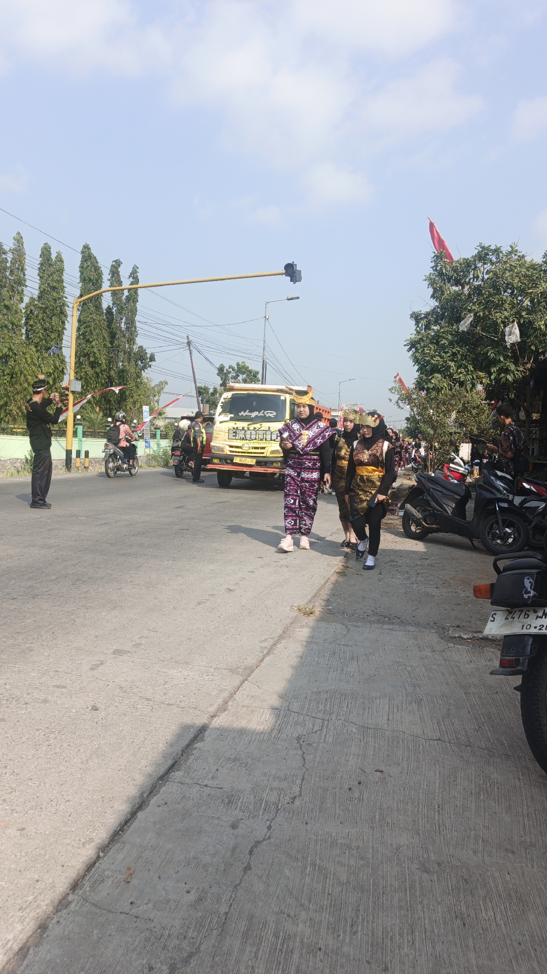 The image depicts a street scene in Sumengko, Indonesia, under a clear sky with scattered clouds. In the foreground, a paved road is visible, with a sidewalk to the right. Several people are present, some walking on the sidewalk and others on the road.
A group of women in traditional attire are walking on the sidewalk. The woman in the front is wearing a purple and white patterned outfit, a headscarf, and pink sneakers. Behind her, two other women are also in traditional clothing, one wearing a gold and brown vest over a black outfit and headscarf, and the other in a similar outfit.
On the road, a yellow truck with the word "EXOTICA" visible on its side is parked. A man in a black uniform and hat is standing on the left side of the road, appearing to be directing traffic or observing. Several motorcycles are parked along the sidewalk and on the road. In the background, trees and buildings are visible, along with power lines and a traffic signal. Red and white flags are also seen decorating the area. The overall impression is of a public gathering or event taking place on a street.