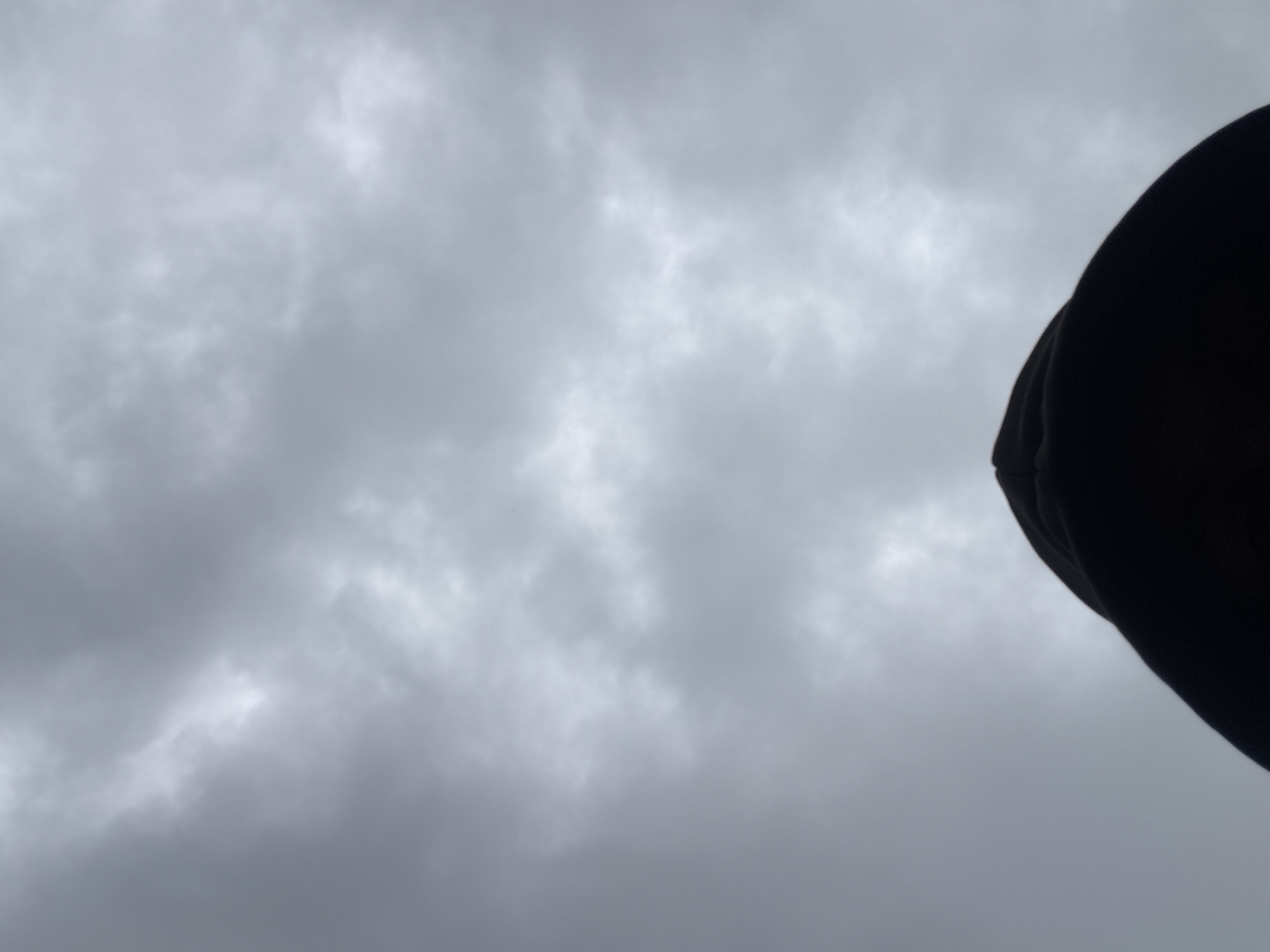 The image shows a person's head wearing a dark hoodie, with the camera pointed upwards towards a cloudy sky. The clouds are grey and overcast, with some lighter patches suggesting sunlight trying to break through. The perspective suggests the person is looking up at the sky. The scene is likely outdoors in Uyo, Nigeria, given the overcast weather common in the region.
