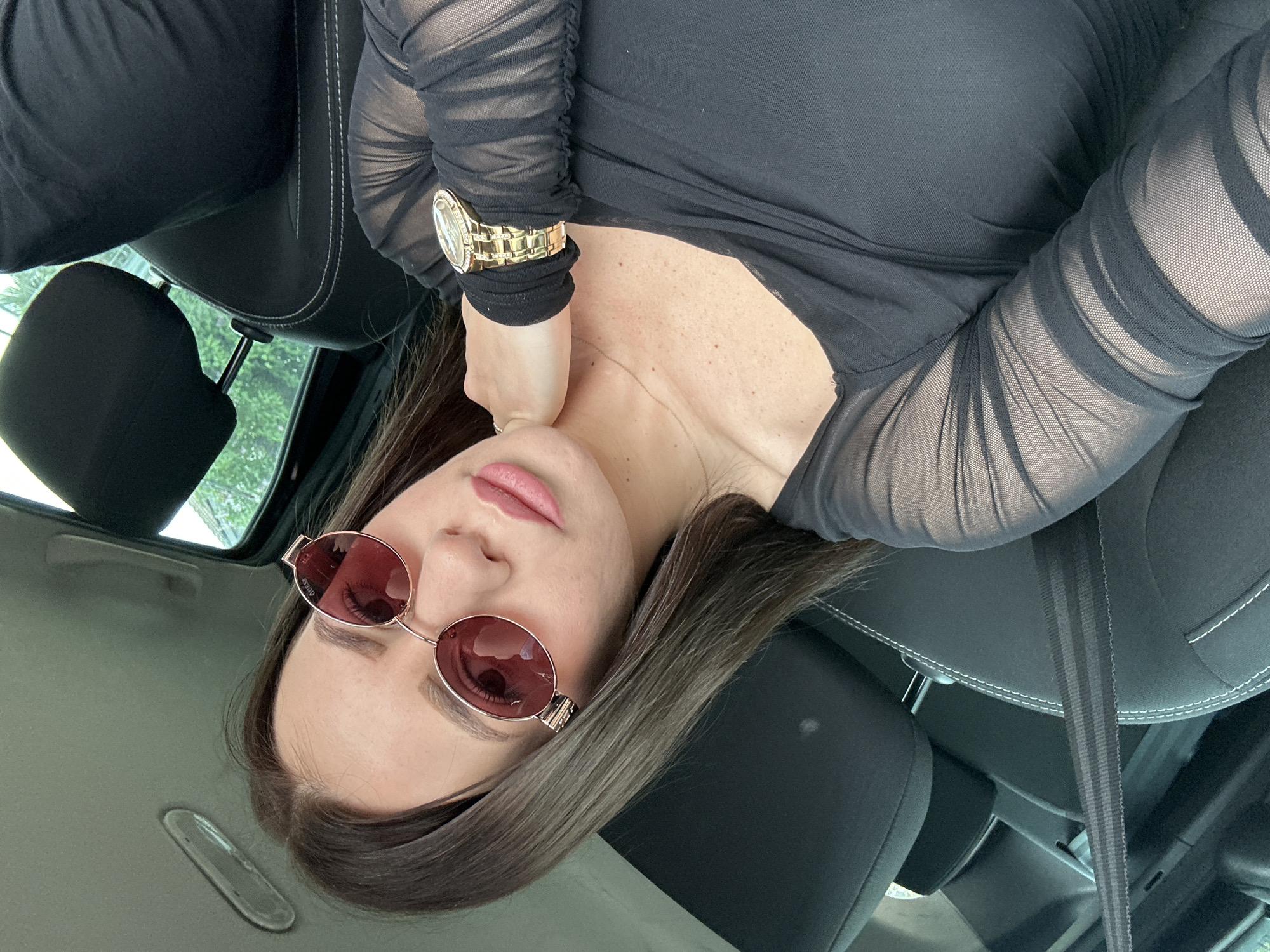 A woman is seated in the driver's seat of a car. She wears a black long-sleeved mesh top, a gold watch, and rose-tinted sunglasses. The car's interior is black leather with white stitching. The environment includes car seats, a seatbelt, and a partial view of greenery outside the window. There is no evidence in the image to pinpoint the exact location in Plovdiv, Bulgaria.