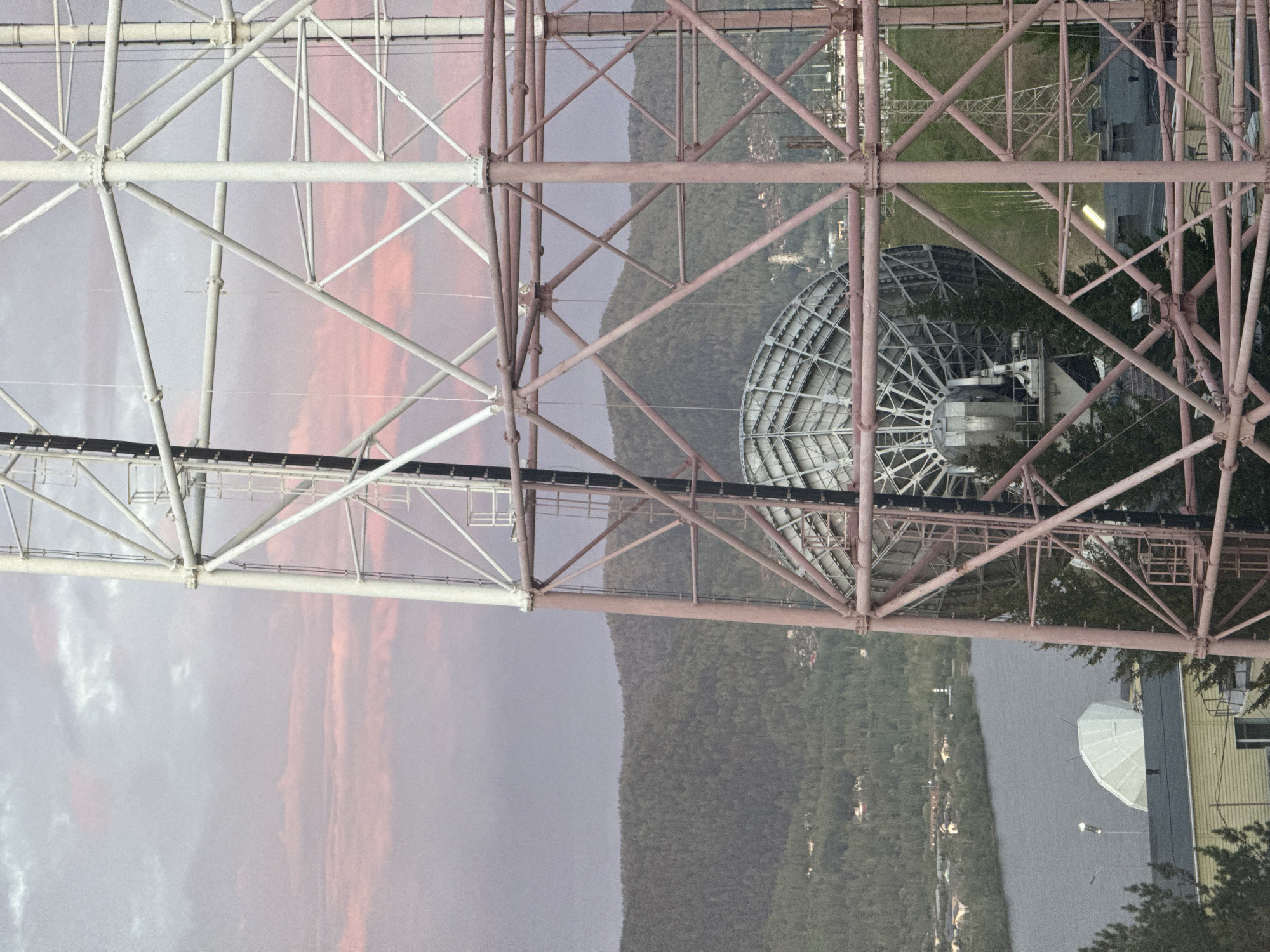 The image shows a structure with a latticework of metal beams and supports, likely an industrial tower or power transmission structure. A satellite dish is visible within the frame of the structure, partially obscured by trees and other built elements. In the background, there is a landscape with a river, a wooded hillside, and distant buildings. The sky is visible through the latticework, with streaks of orange suggesting sunset or sunrise. This is a view of the Krasnoyarsk TV tower from the ground, located at Nikolayevsky Prospekt, 16а, Krasnoyarsk, Russia, 660021.