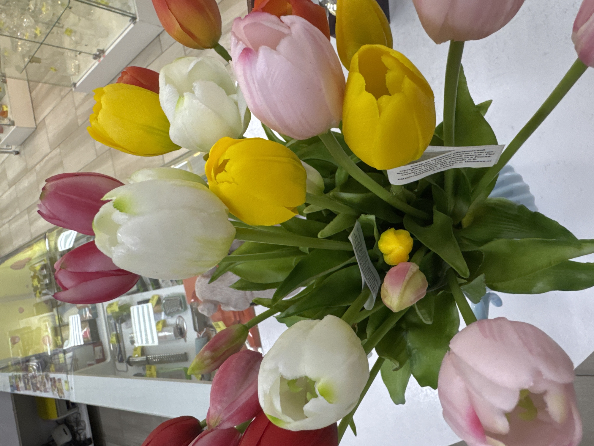 The image shows a bouquet of artificial tulips in various colors, including yellow, white, pink, and orange. The flowers are arranged in a vase. The background appears to be a store display case, likely in a shop or commercial setting in Klin, Russia, though the exact location within the city is not identifiable. The case contains various items for sale, and a mirrored surface reflects the upper portion of the bouquet.