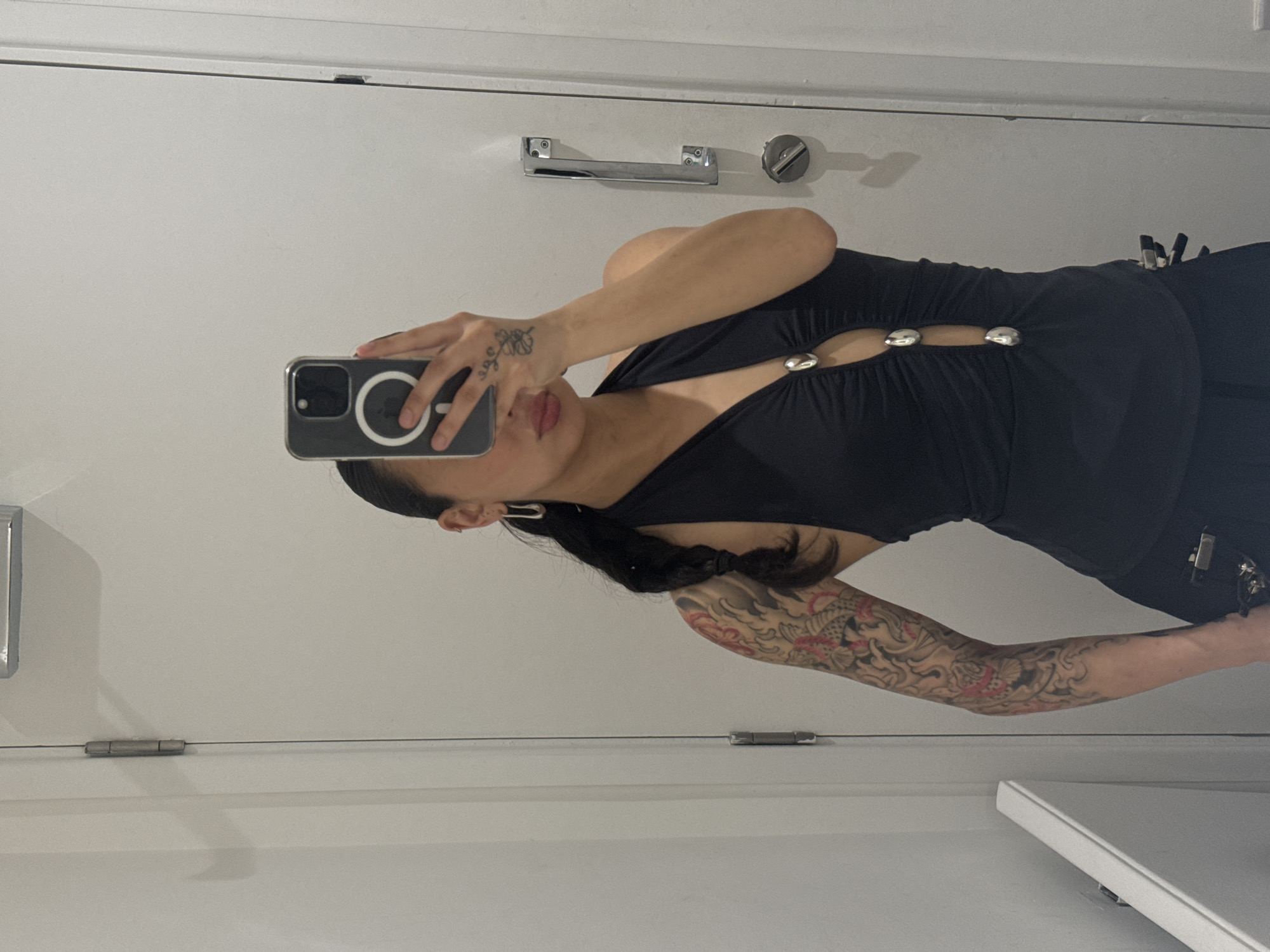 A woman is taking a selfie in a bathroom. She is wearing a black top with metallic buttons. The bathroom has a white door with a metallic handle and hinges. The location is not discernible as being in London.