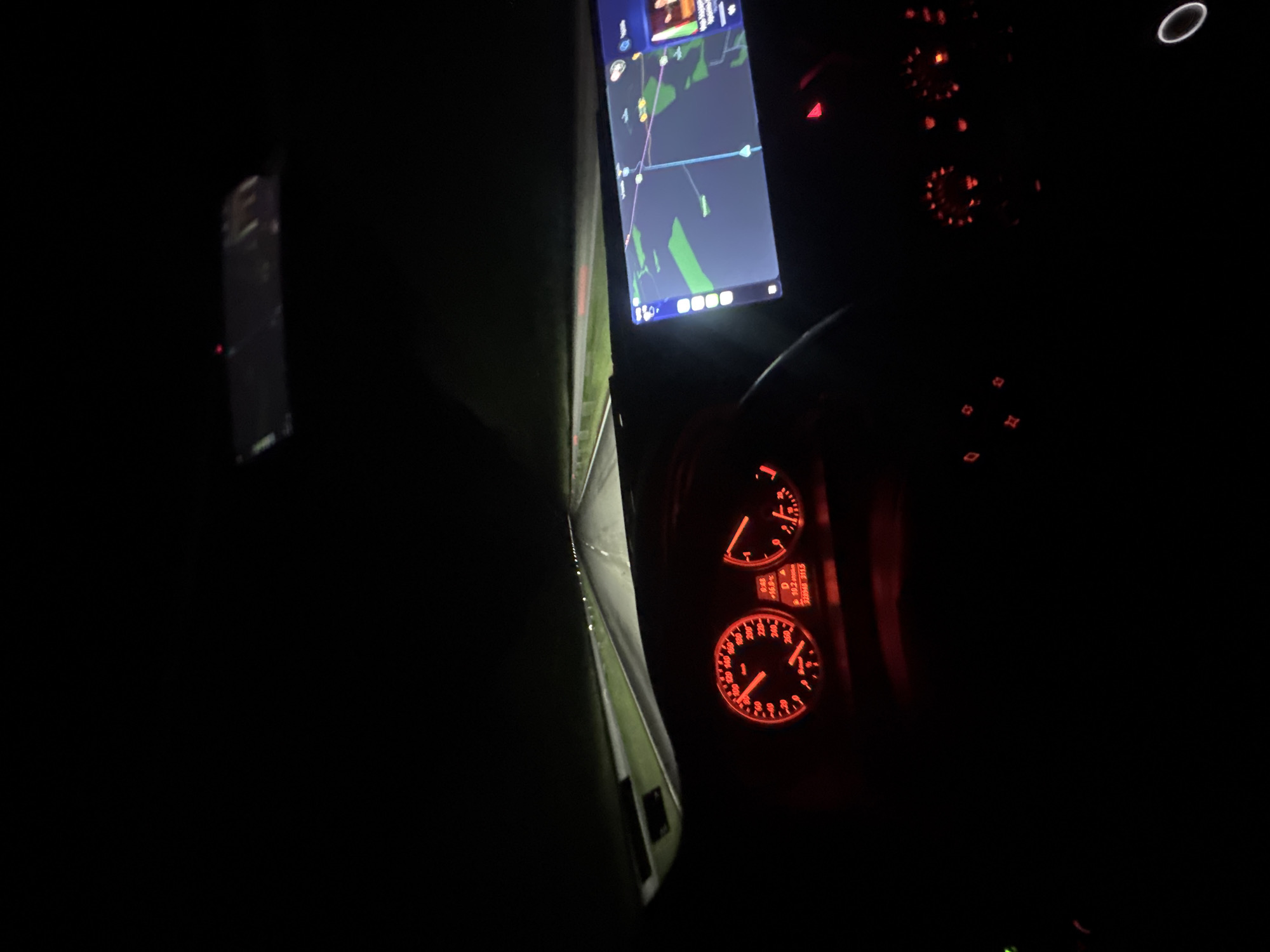 The image, taken from the driver's perspective of a vehicle moving at night, displays a dashboard and a navigation screen. The navigation screen shows a route with green and blue details, suggesting geographical features and roads. The dashboard is illuminated in red, showcasing the speedometer and other gauges, with the speedometer reading indicating a moderate speed. The text on the odometer reads "328948 311.5" and indicates 10.2 kilometers, 16% fuel, and 48 minutes. There is an abstract reflection on the driver's side window showing green trees and road. Based on the available information, there is no information to identify the specific location in the city of Mogila, Bulgaria.