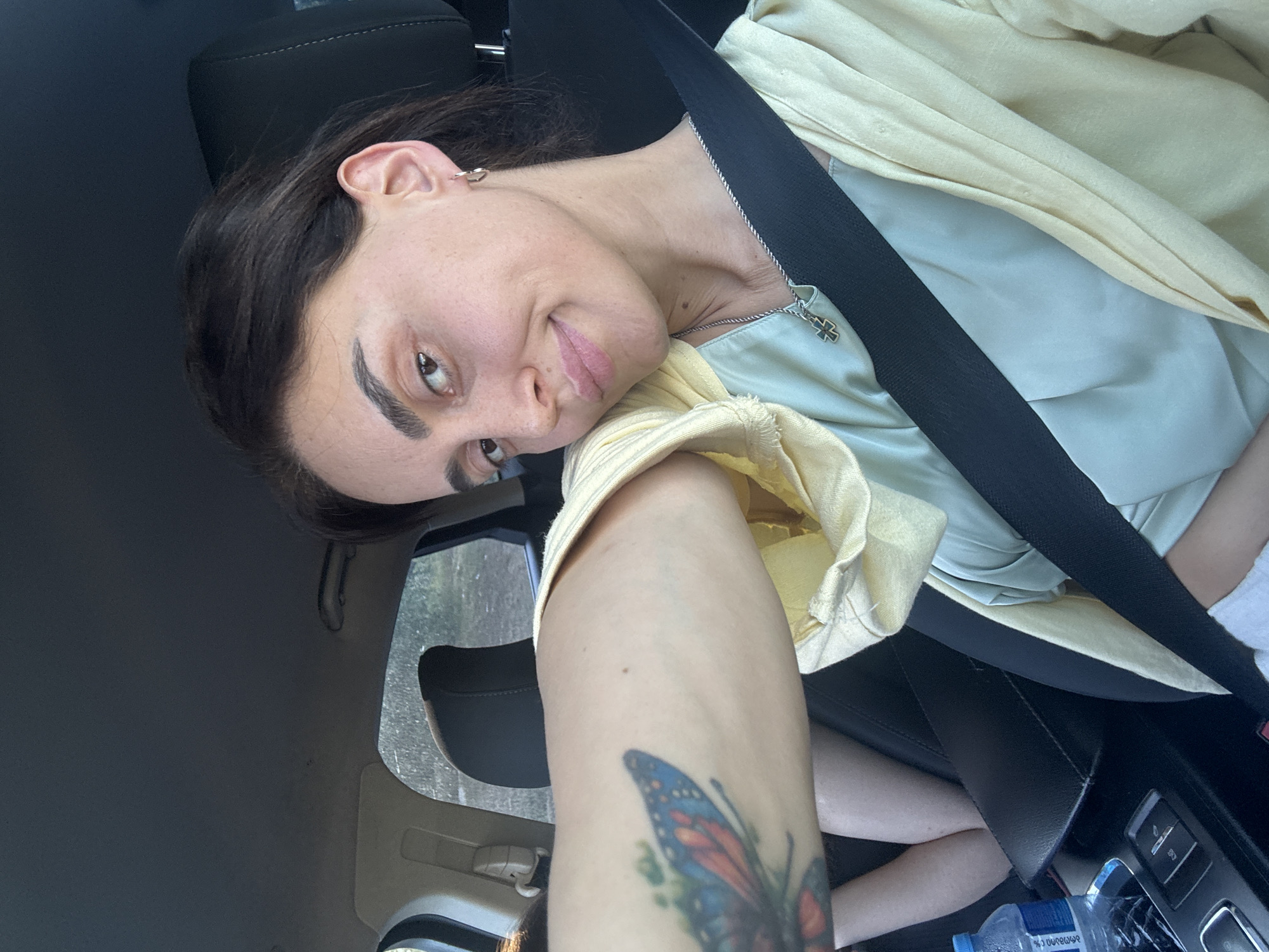 A woman with dark hair, wearing a light green shirt and a yellow jacket, is seated in the passenger seat of a car. She is wearing a seatbelt, a silver necklace with a cross pendant, and hoop earrings. A tattoo of a butterfly is visible on her left arm. A bottle of water is on the center console. The car is moving through a tree-lined area that appears to be on the outskirts of Tbilisi, Georgia.