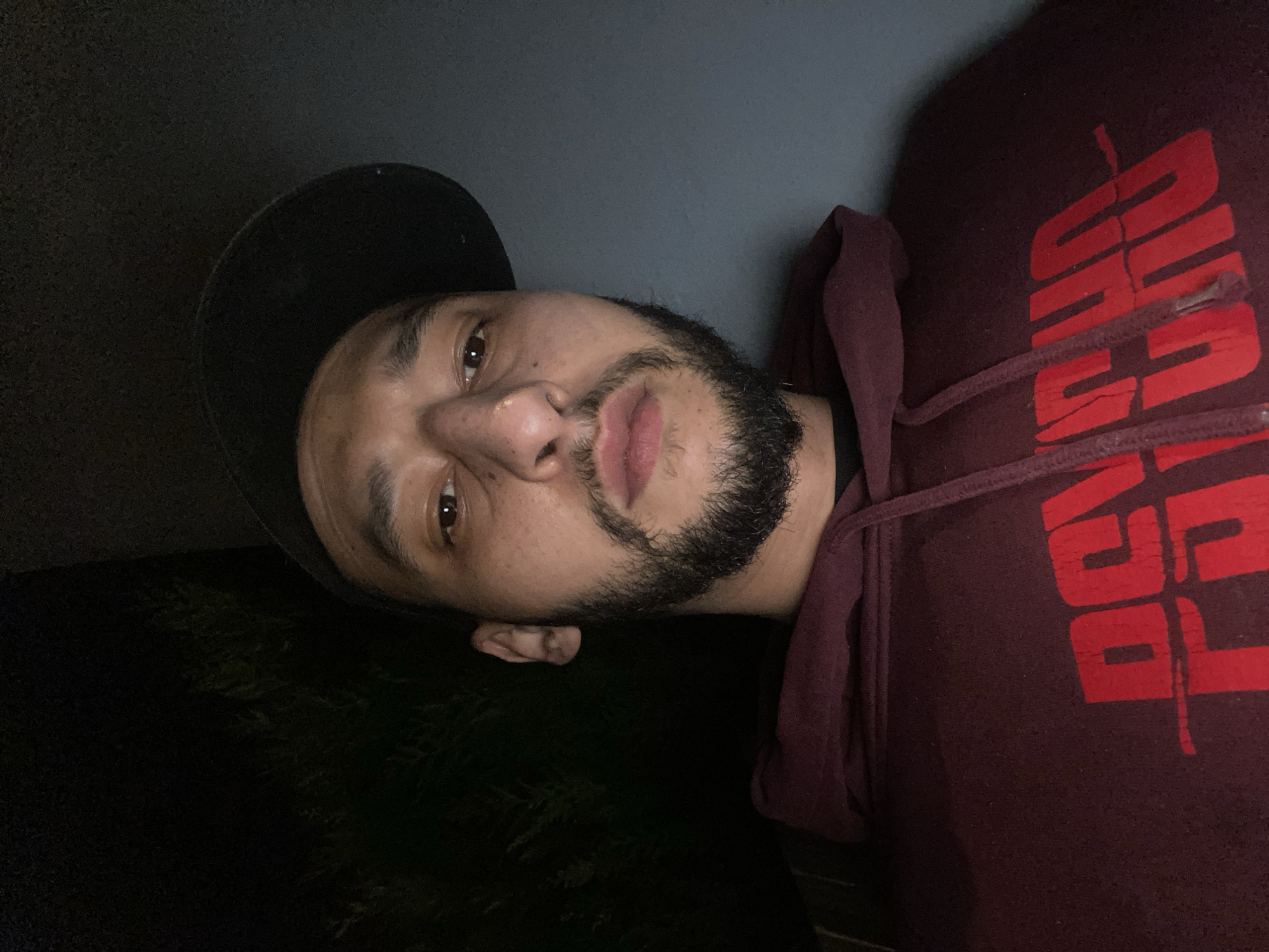 A person is pictured wearing a black hat and a maroon hooded sweatshirt with a red logo. The person has a beard and a nose ring. The setting appears to be dimly lit, with what may be foliage visible in the lower left. The environment's location in Schiltigheim, France, is not evident.