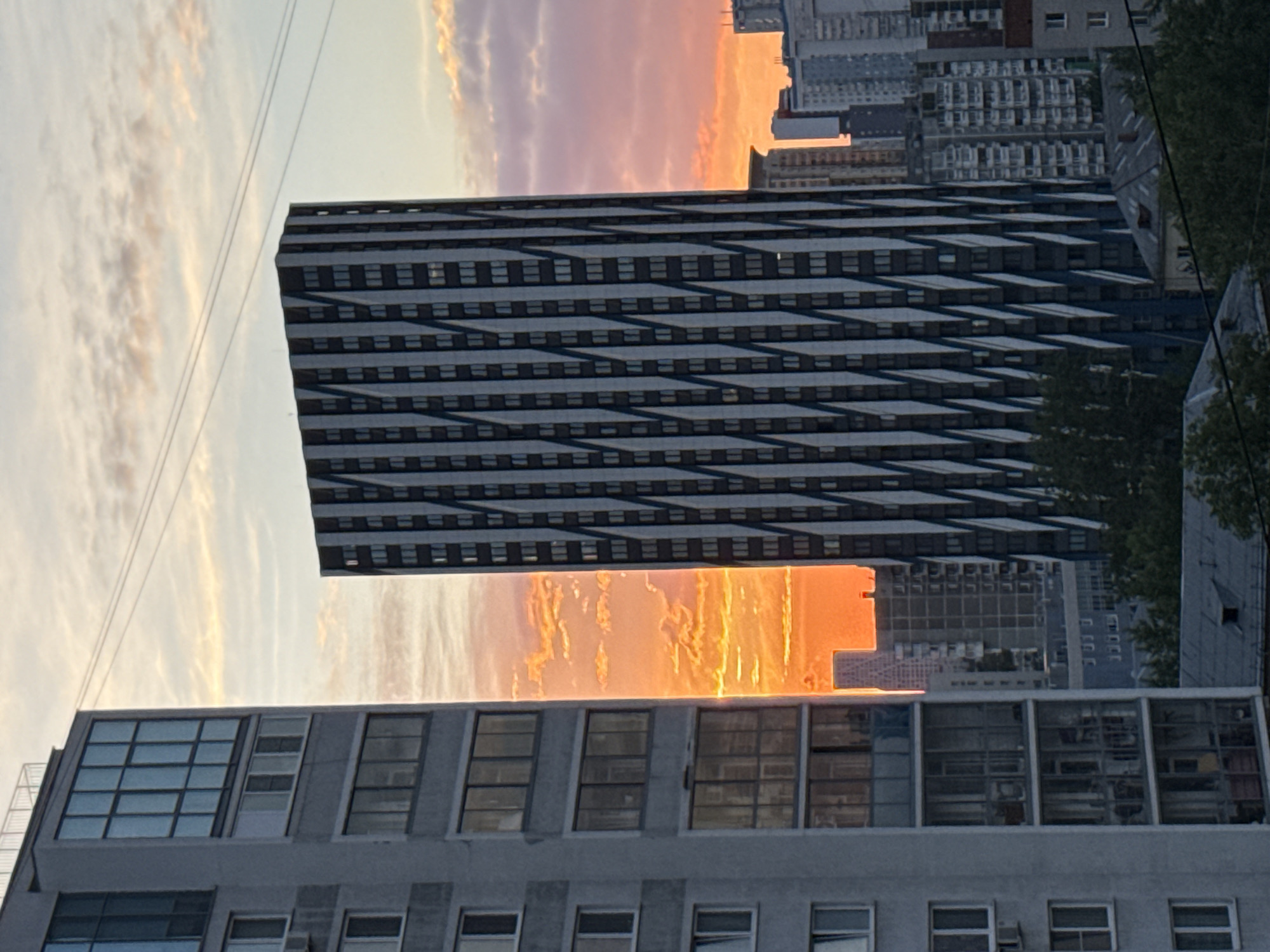The image depicts an urban scene in Yekaterinburg, Russia, featuring multiple buildings against a colorful sky. The foreground building is a light-gray concrete structure with a repeating pattern of windows, some of which have small air conditioning units. Above it, there is a taller, modern building with a striped black and white pattern. An intense orange and yellow sunset appears in the sky between the buildings, suggesting the setting sun. Further back are other high-rise buildings, hinting at a densely populated urban environment. Power lines are visible in the top left corner of the image. The foliage of trees can be seen on the right side of the image.