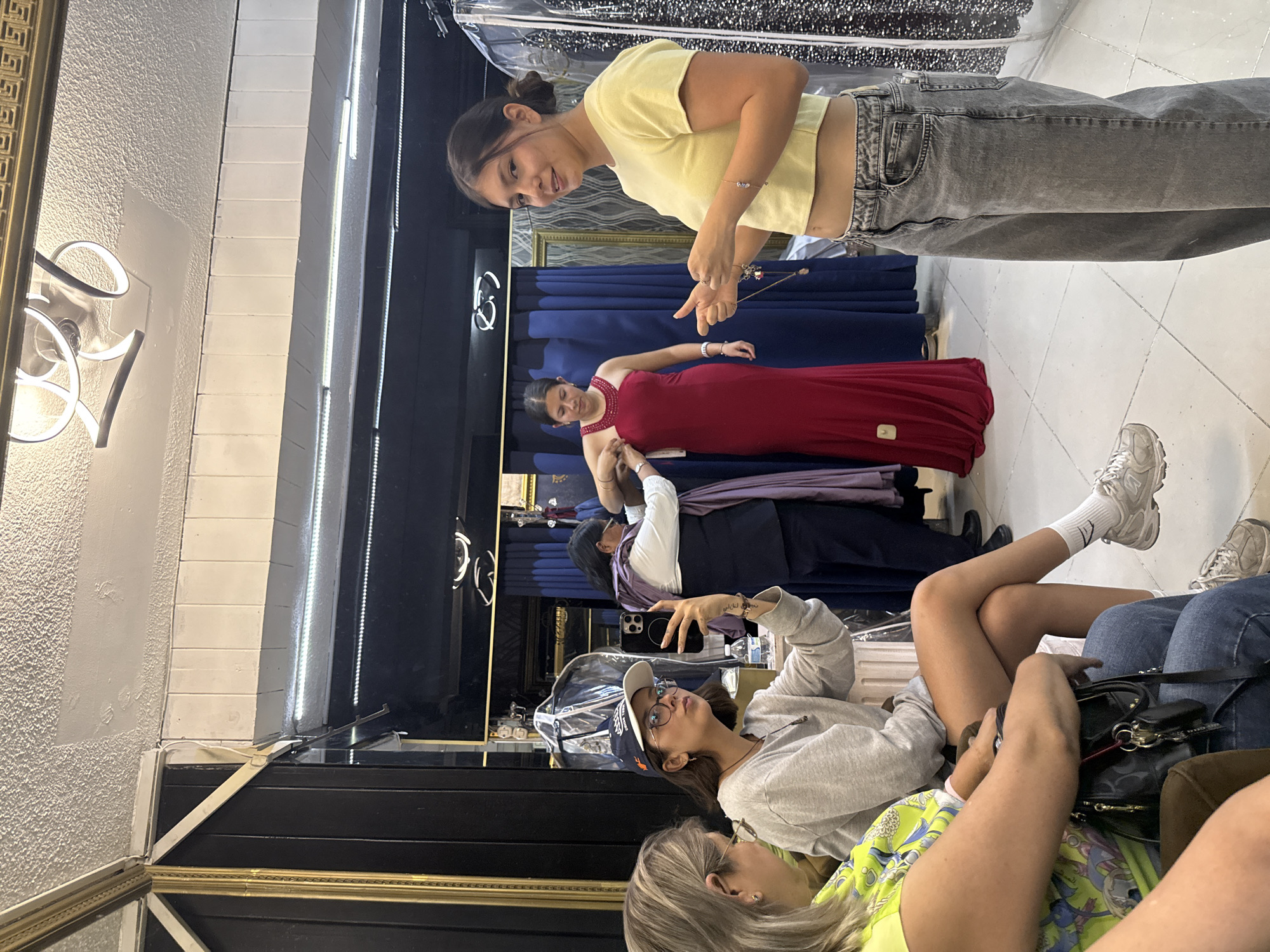 The scene appears to be inside a dress shop in Puebla, Mexico. A young woman is trying on a long red dress while another woman, wearing a yellow shirt and gray jeans, stands nearby, appearing to offer assistance or advice. An older woman in a white shirt and dark pants appears to be assisting with the dress fitting. In the foreground, other women are seated, possibly waiting. One is wearing a baseball cap and gray sweatshirt, and another is wearing a yellow shirt with a patterned print. A black handbag is present in the foreground. The environment includes dressing rooms with blue curtains and mirrored walls. A modern light fixture is visible on the left.
