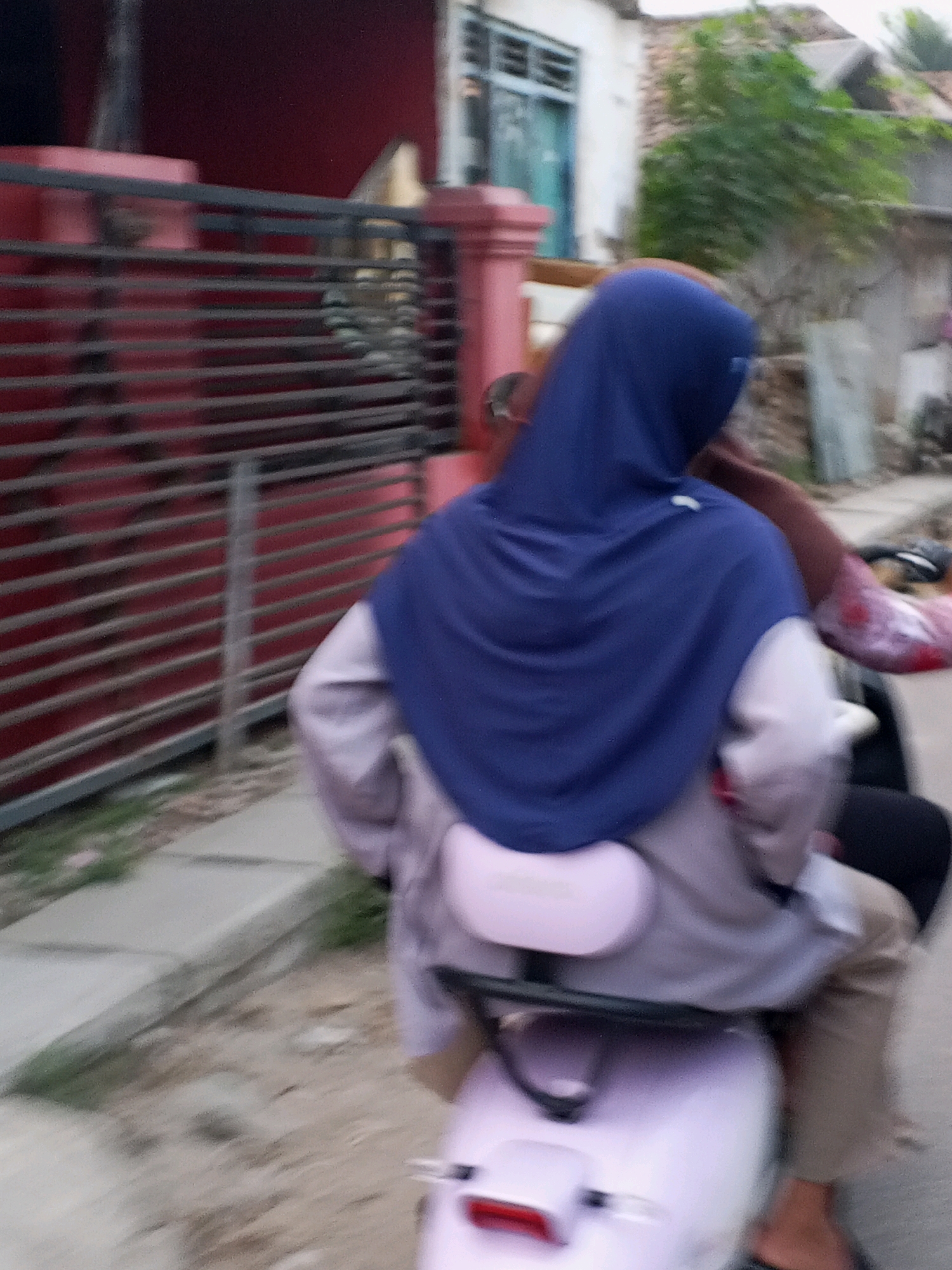 A slightly blurry, eye-level shot captures two individuals on a light purple scooter traveling down a paved street in Kadikaran, Indonesia. The person in front is wearing a dark blue hijab, a light gray shirt, and tan pants. They are seated on the scooter. Behind them, another person, partially visible, appears to be leaning forward. The scooter has a light pink backrest. To the left of the street is a red fence with horizontal bars, and beyond that, a dark red building. To the right, there are houses with white walls and green foliage. The ground is a mix of dirt and paved surfaces, with some small green plants growing near the curb. The overall impression is of everyday movement through a residential area.