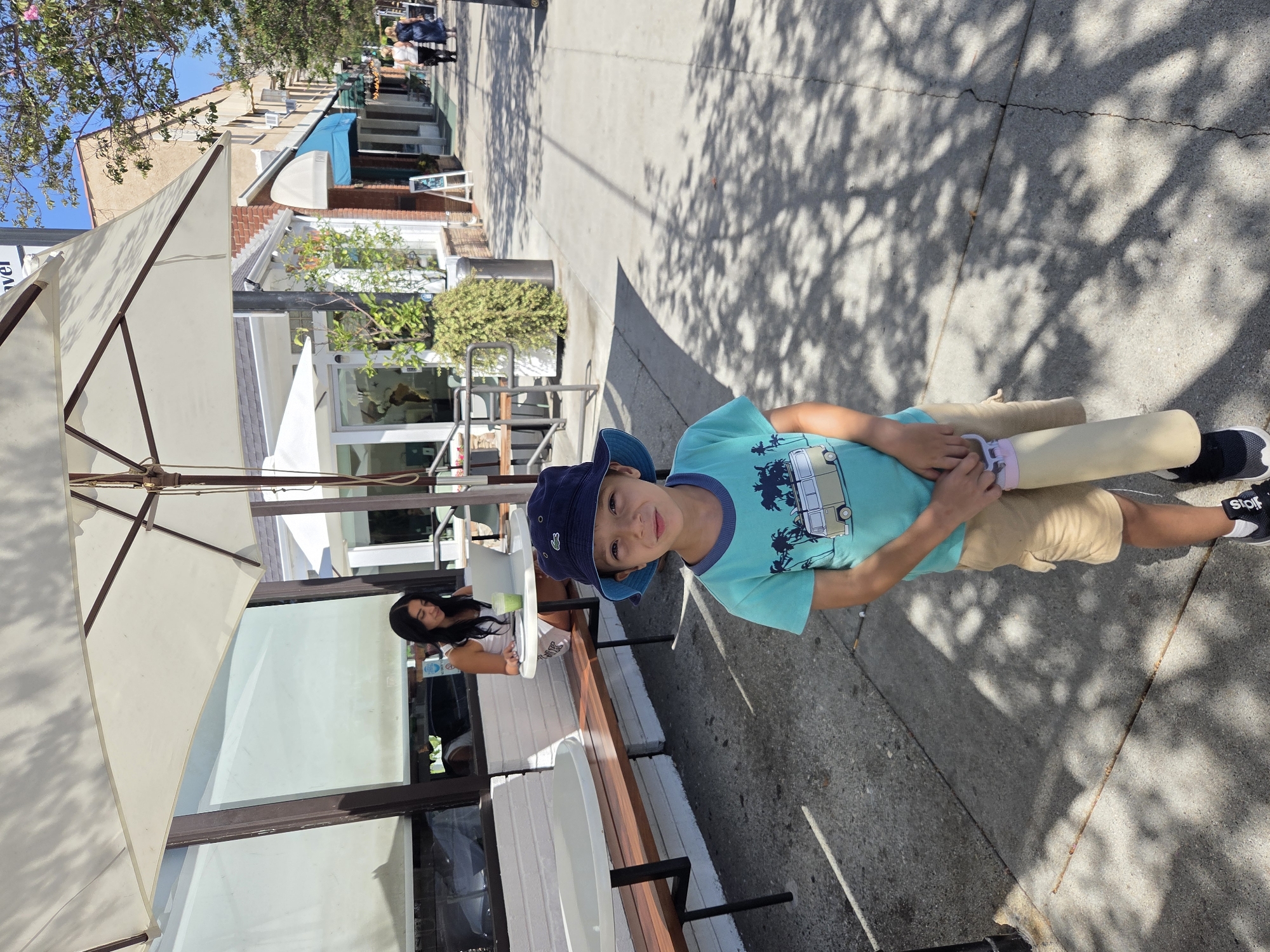 A young boy wearing a blue bucket hat and a turquoise t-shirt with a graphic of a van and palm trees, paired with khaki shorts, stands on a sidewalk. He holds a light-colored water bottle in his right hand. Behind him, a woman with dark hair is seated at a table outdoors, looking at a laptop. The scene appears to be in front of a cafe or restaurant, with outdoor seating and a large umbrella providing shade. The sidewalk is paved, with dappled shadows from trees overhead. In the background, buildings line the street, suggesting an urban or commercial district. This image was taken in Glendale, California, United States of America.