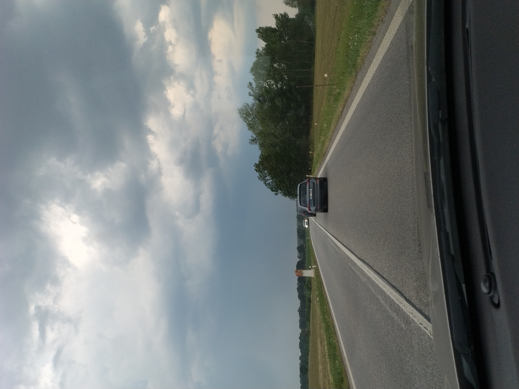 The image depicts a view from inside a vehicle traveling along a road in Plástovice, Czechia. The scene is dominated by a dramatic sky filled with dark, heavy clouds, suggesting impending or ongoing rain. The road is paved and marked with a white line, indicating it's a two-lane highway. To the right of the road, there is a grassy verge with scattered wildflowers and a line of trees forming a dense forest border.
Several vehicles are visible on the road. In the foreground, a dark-colored SUV is driving ahead, its taillights illuminated. Further down the road, another vehicle, possibly a car, is also visible. The perspective suggests the photo was taken from the passenger seat or driver's seat of a moving vehicle, as evidenced by the dark interior frame on the right side of the image.
Along the roadside, there are a few small, white rectangular markers, possibly mile markers or signs, and a distinctive orange and white structure. The overall mood is slightly somber due to the overcast sky, but the presence of the road and other vehicles implies normal travel or commuting. The time of day appears to be daytime, though the cloudy conditions obscure direct sunlight. No people are clearly identifiable within the image. The license plate of the SUV is partially visible, displaying characters that appear to be "E4R 7569".