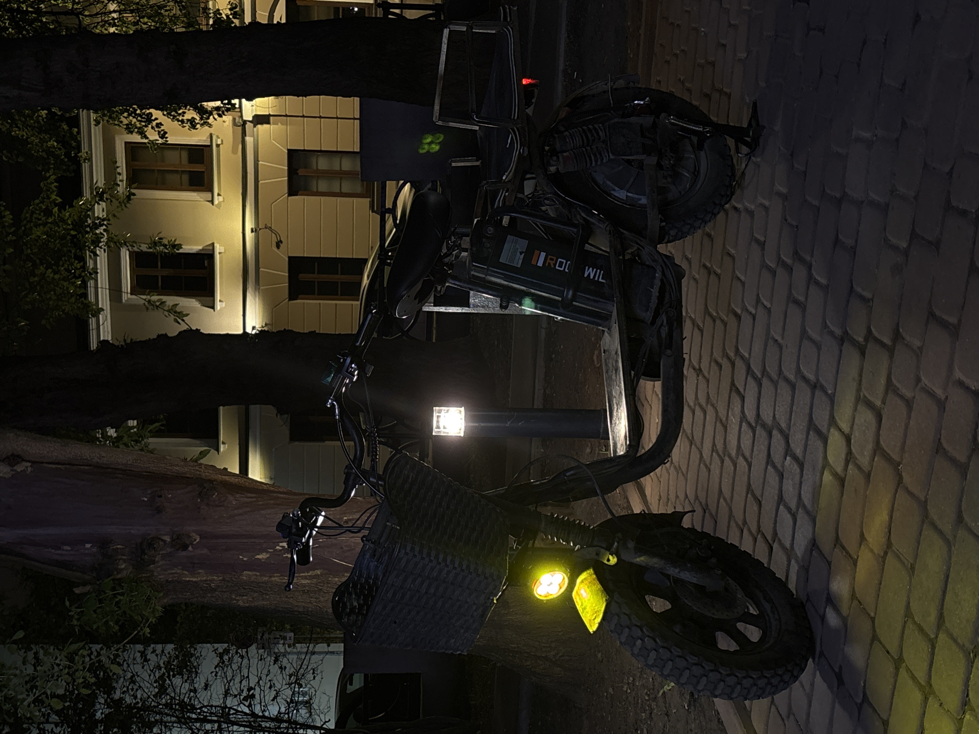The image shows an electric moped parked on a brick-paved area at night. In the background, there's a building with multiple windows and a tree. The moped has a basket on the front and illuminated yellow lights. The moped's brand, "RODWIL", is visible. A street light illuminates the scene. Given the style of the building, the bricks, and architecture, the location is likely in a historical district of Moscow, Russia, though specific address details are not discernable from the image.