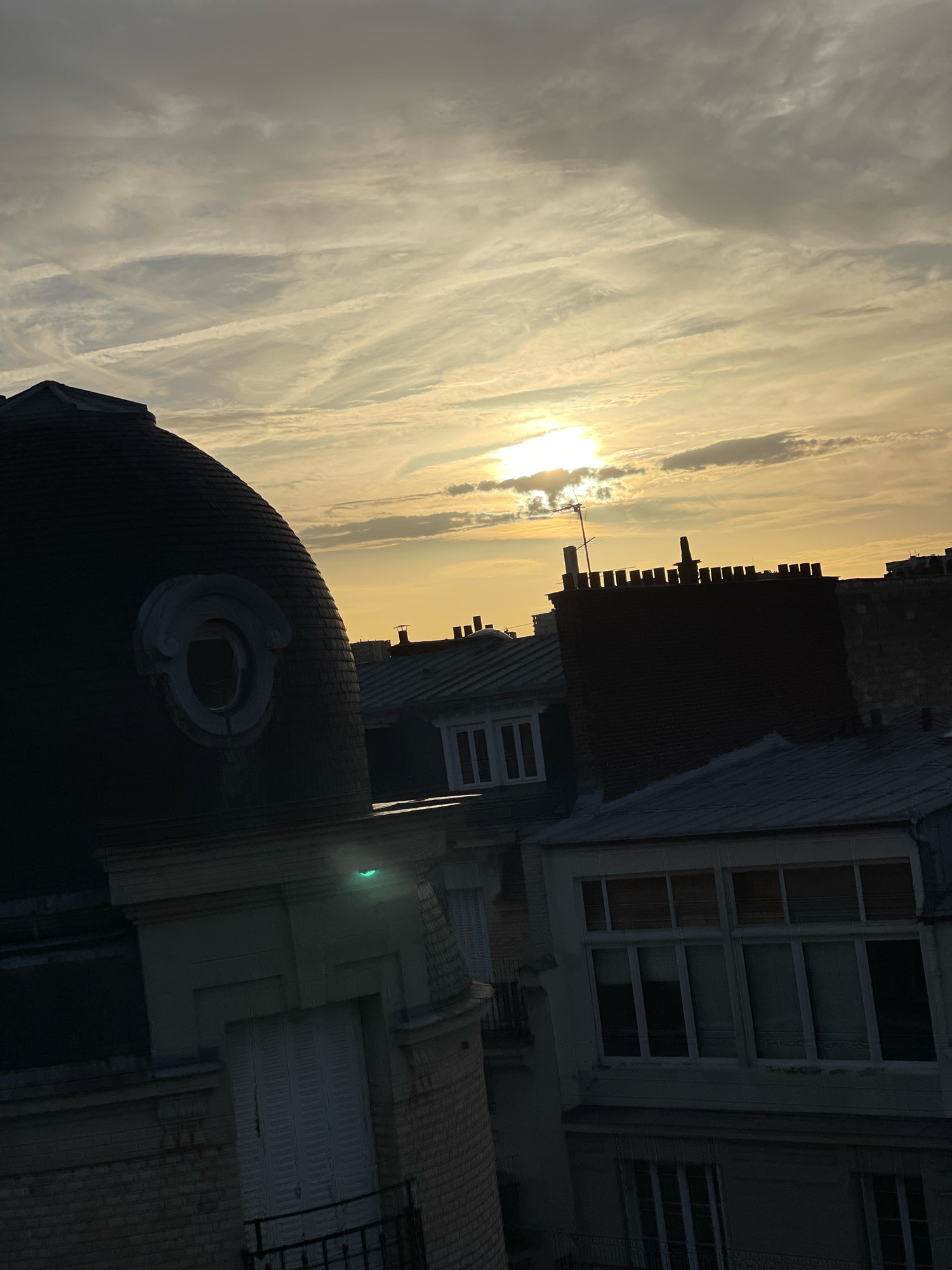 The image depicts a view of Parisian rooftops at sunset. The sky is filled with a mix of light and dark clouds, with the sun peeking through, casting a warm, golden glow over the scene. Several buildings with characteristic Parisian architecture are visible, including a domed structure with an ornate circular window on the left, a dark brick building with a crenelated top, and other buildings with sloped roofs and multiple windows. The rooftops are varied in material and design, typical of the urban landscape of Paris. The overall impression is one of a tranquil evening in the city, with the architecture and natural light creating a picturesque view.