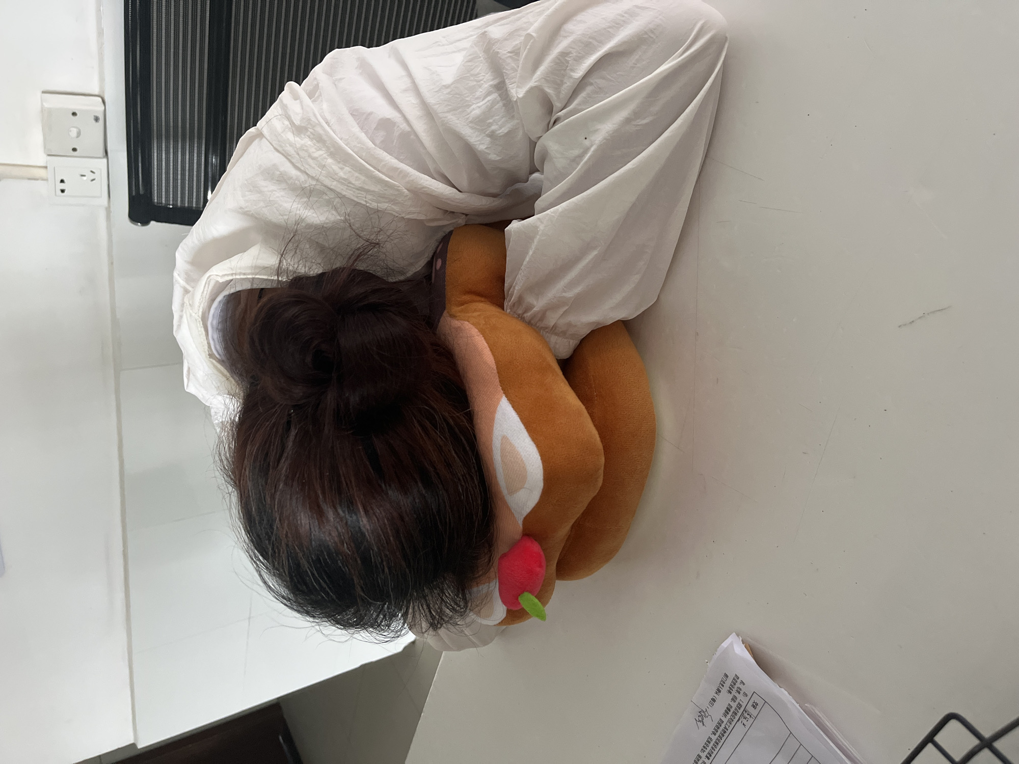A person with dark brown hair pulled into a bun is resting their head on a plush toy shaped like a reindeer. The person is wearing a white outfit with a wrinkled texture. The scene appears to be indoors, with a white desk surface visible beneath the person and the toy. A paper with Chinese characters and a partially visible office chair with a black mesh back are also present. A power outlet is seen on the wall. The photo was possibly taken in Jiangjin, China, based on the Chinese text on the paper.
