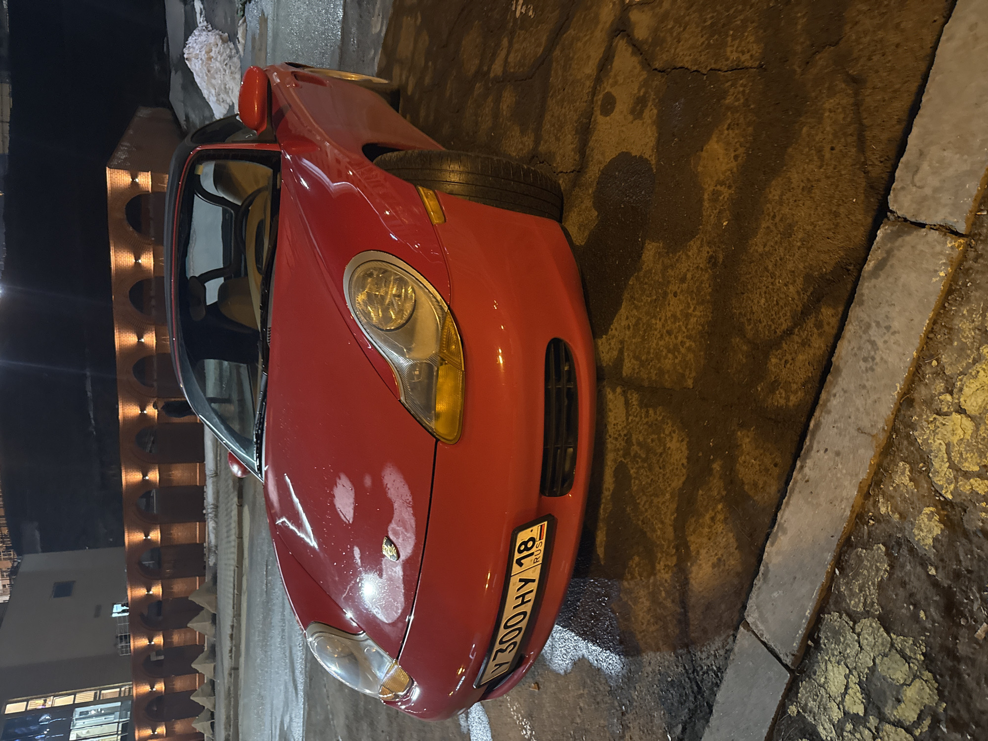 The image shows a red Porsche parked on a wet city street in Izhevsk, Russia. The car's license plate reads "Y 300 HY 18 RUS". The background includes a building with illuminated arches and residential-looking windows. The environment is dimly lit, suggesting it is night. The road appears to be paved and wet. A concrete curb separates the road from a patch of ground that appears to have yellowing vegetation.